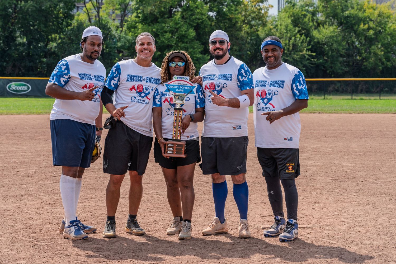 Five charity softball players pose together on the field holding a large trophy after their game.