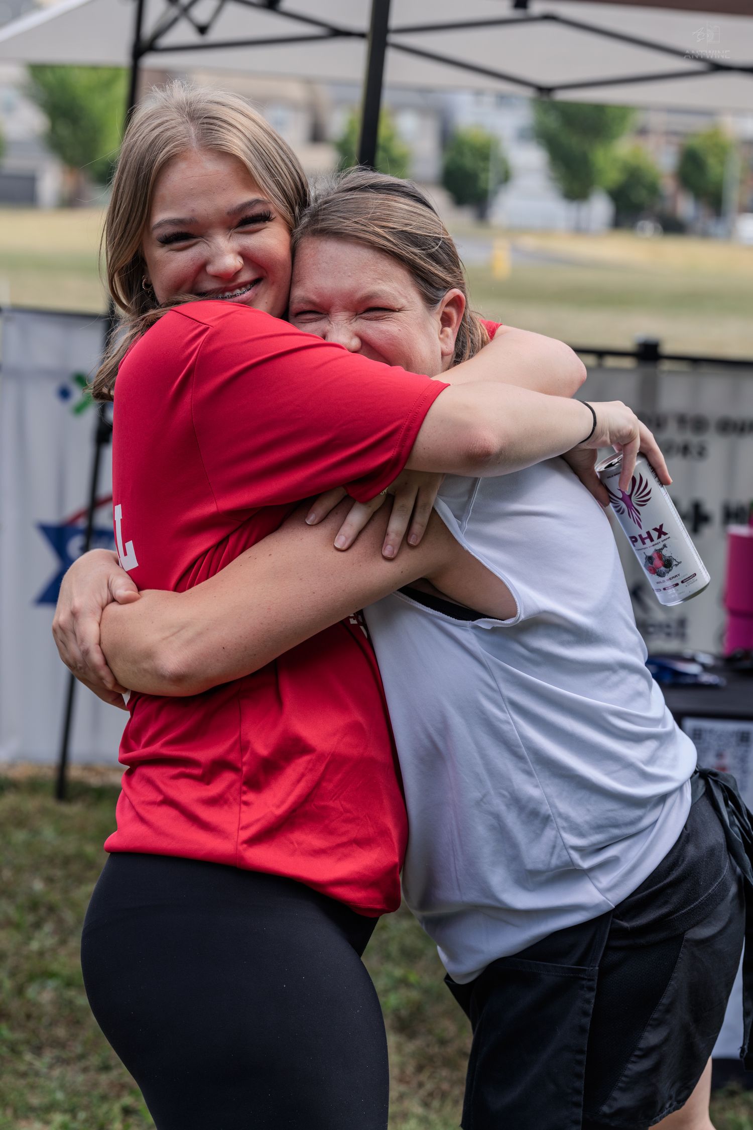 Two women share a warm hug outdoors at a community event, one in red shirt holding a can.