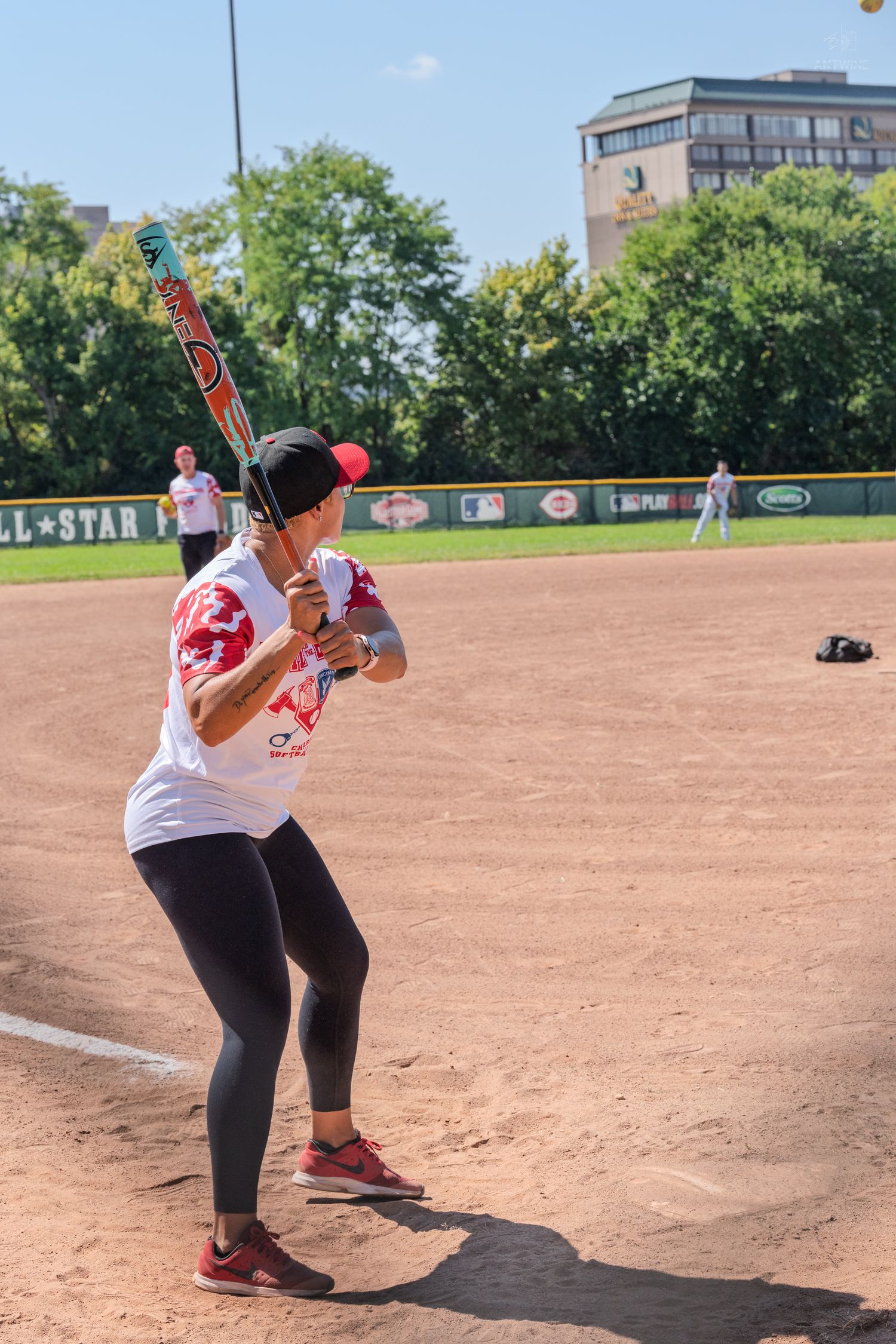 Woman in white shirt and black leggings swinging a bat at a softball game on a sunny day.