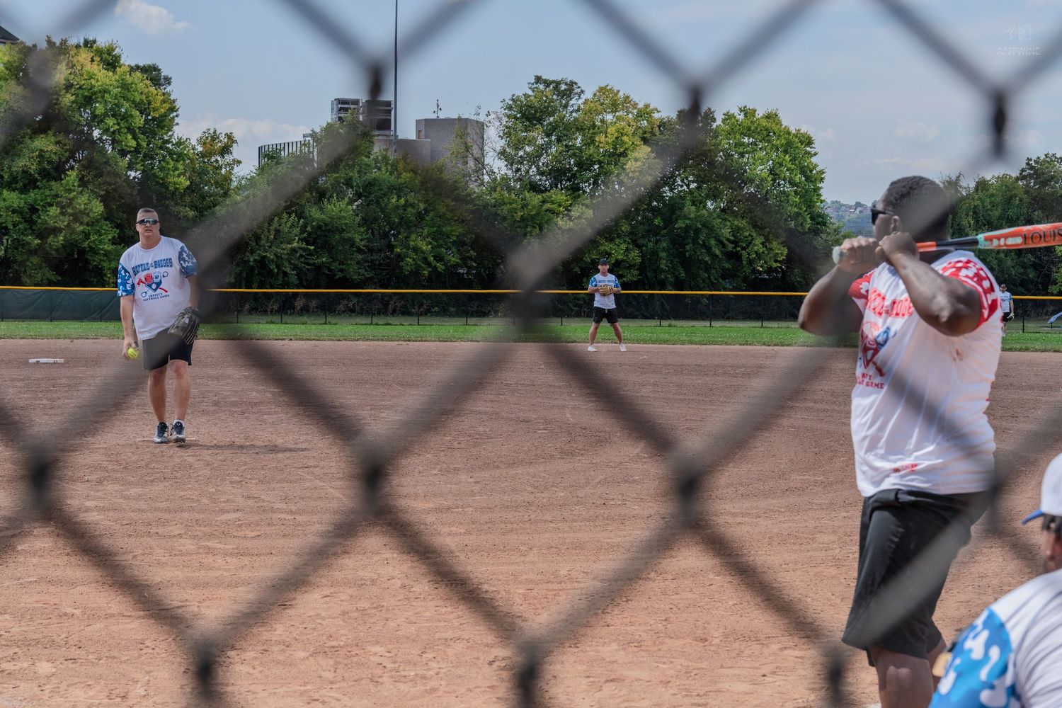 Softball pitcher throwing to a batter viewed through a chain-link fence during an outdoor game.