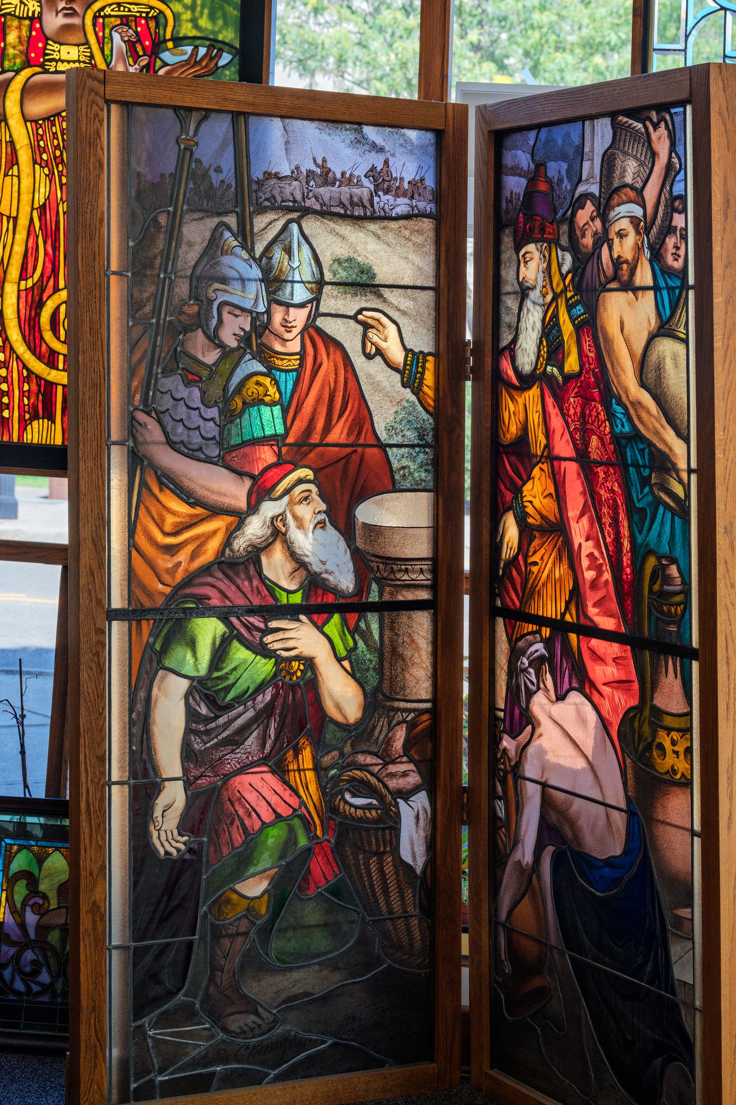 Colorful stained glass window panels depicting biblical scenes with robed figures, soldiers, and elders in vivid reds, blues, and greens.