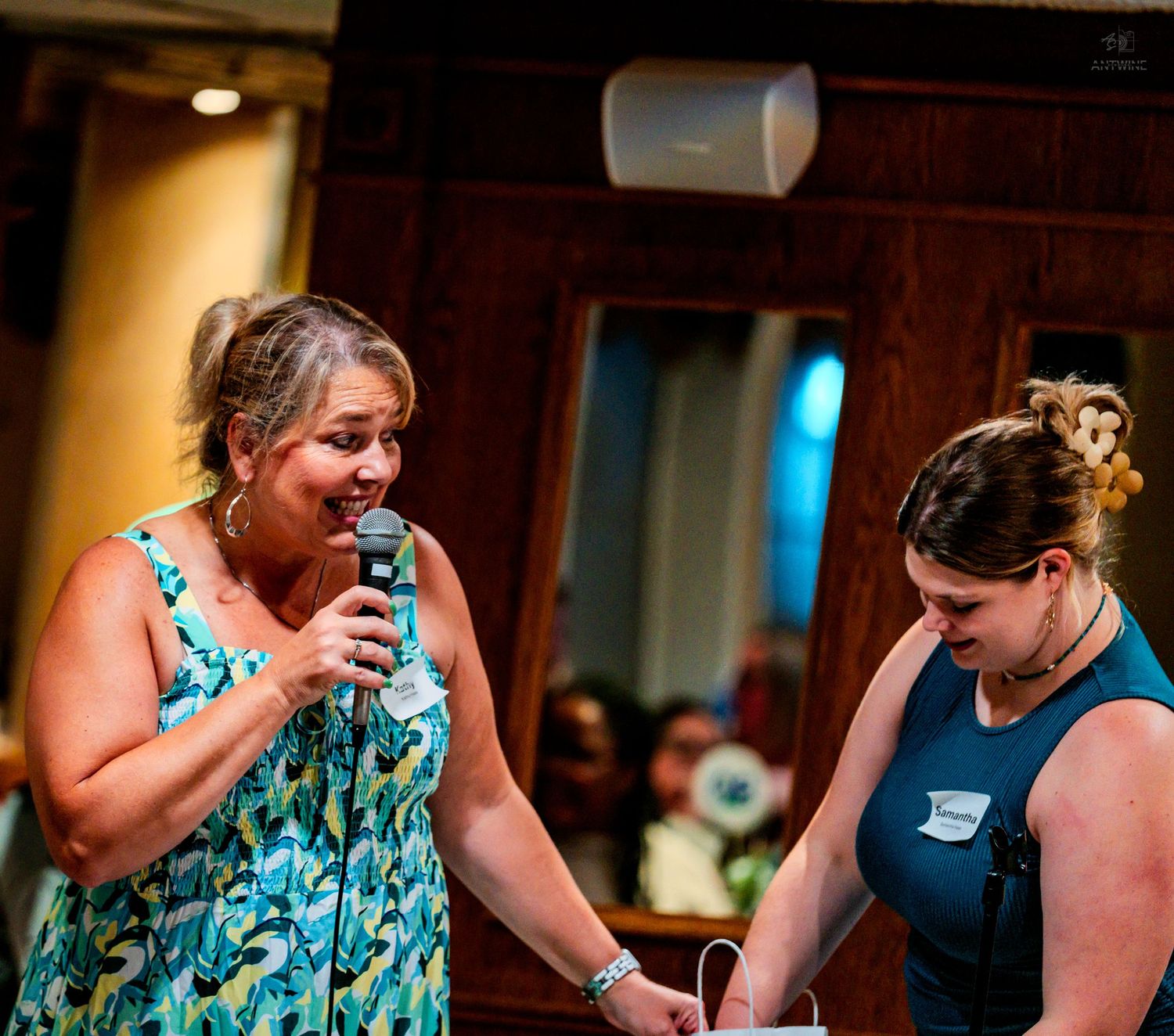 Two women sharing a moment on stage, one speaking into a microphone at an event.