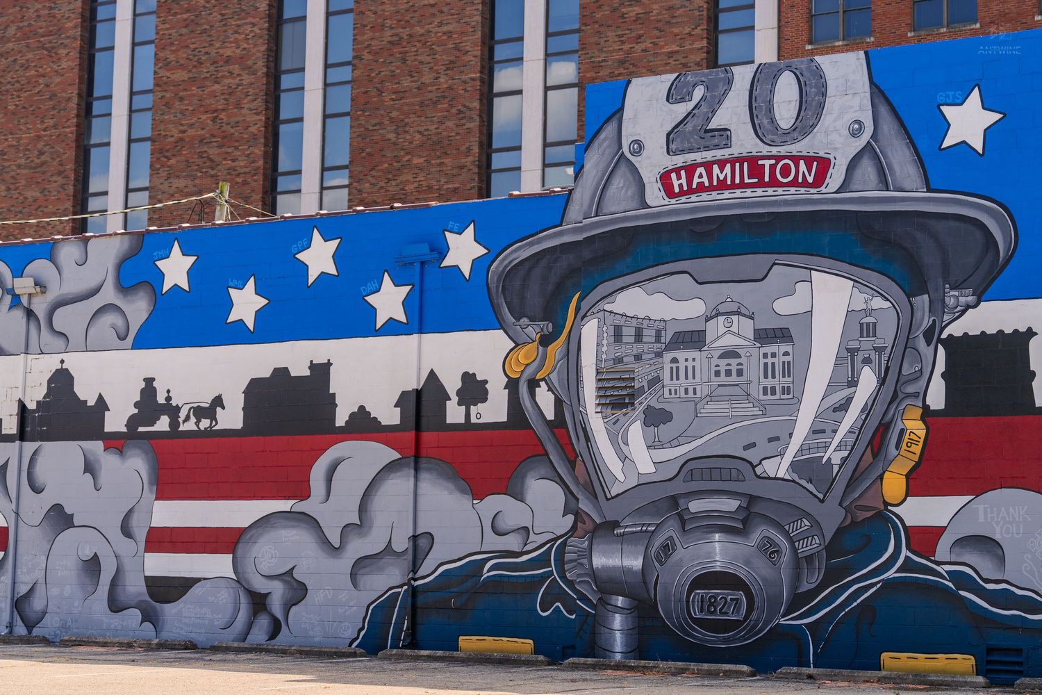 Firefighter mural with American flag, Hamilton Station 20 helmet, and cityscape on brick wall.