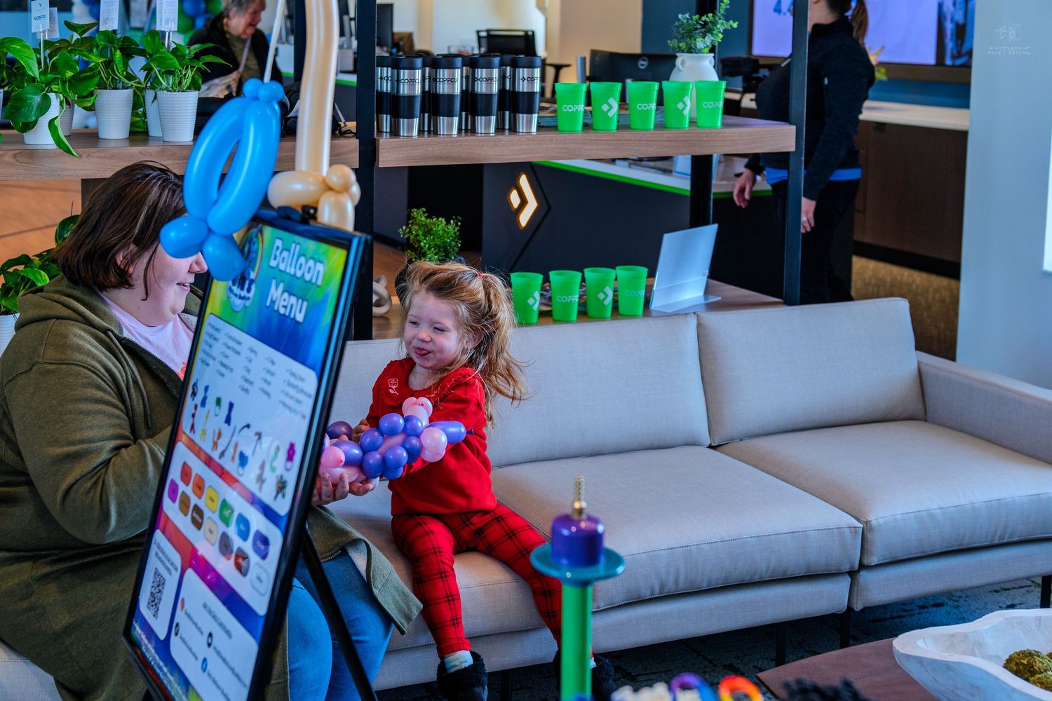 Young girl in red outfit sits on white sofa beside woman reviewing colorful menu display in modern retail store.