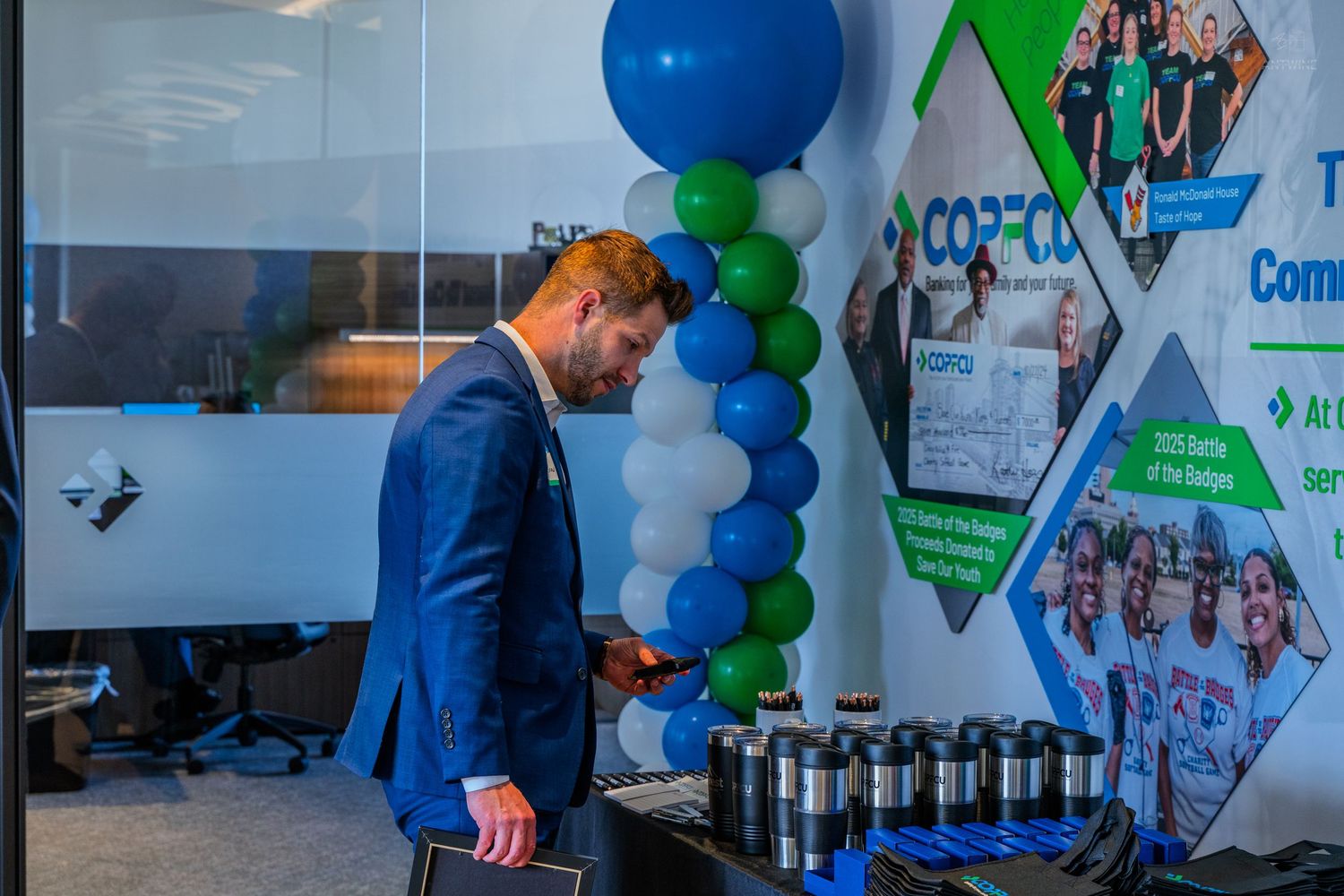 Man in blue suit checks phone near green and blue balloon column at a COPCO corporate event.