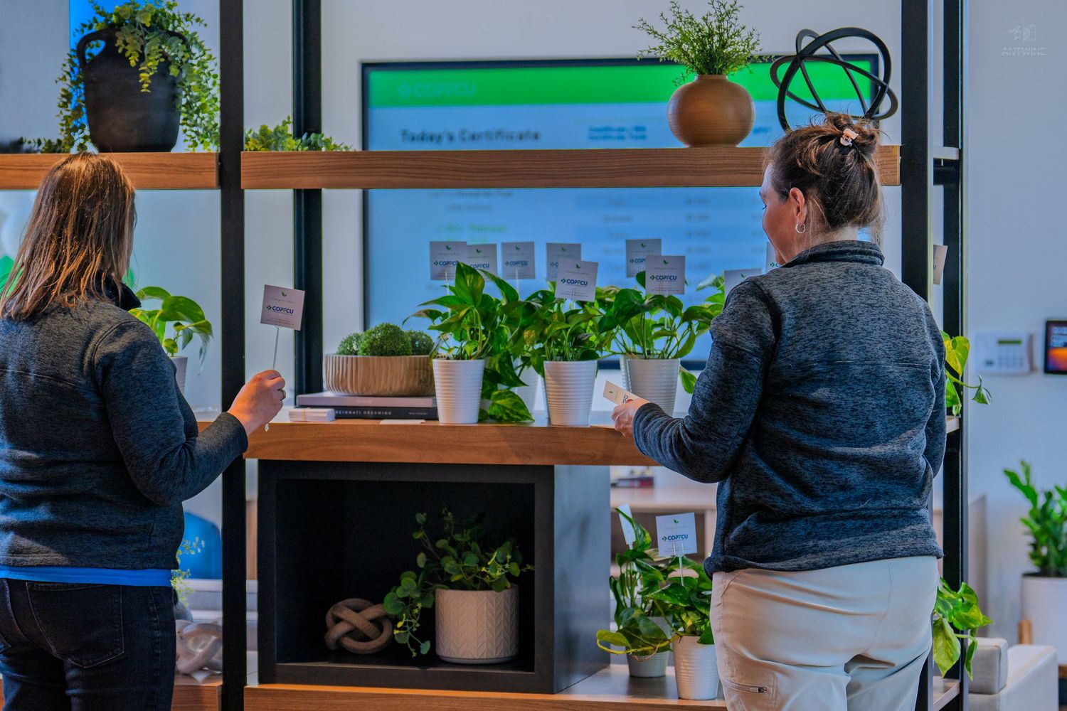 Two women browsing potted plants on wooden shelves near a digital display screen indoors.