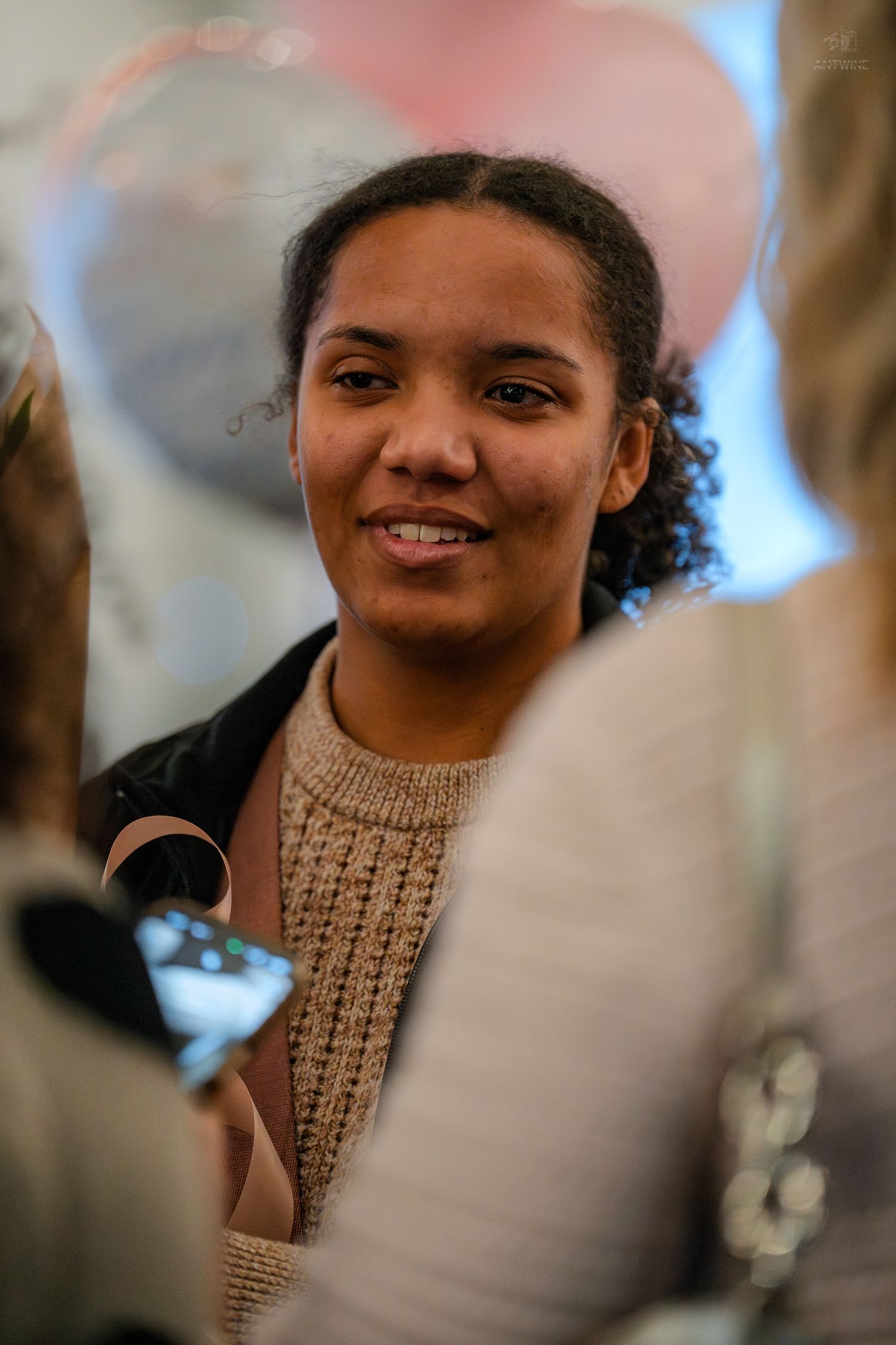 A young woman smiles warmly while surrounded by friends at a celebration with pink balloons in background.