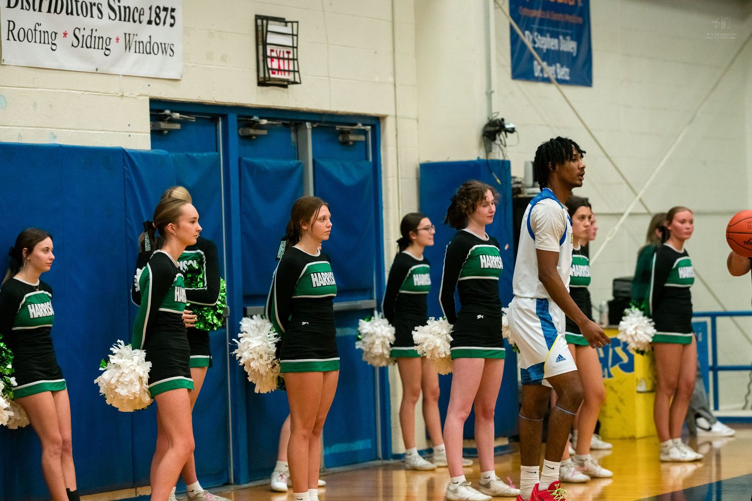 Green uniformed cheerleaders and tall basketball player stand along gymnasium wall during game.