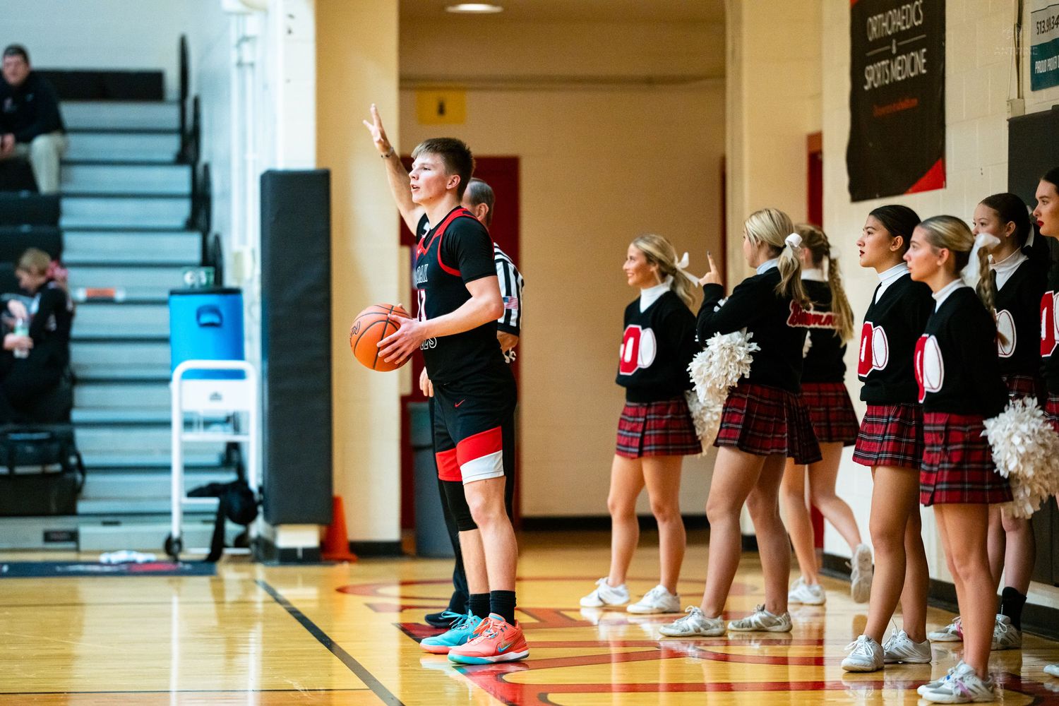 A basketball player in black uniform signals while cheerleaders in plaid skirts line the sideline.