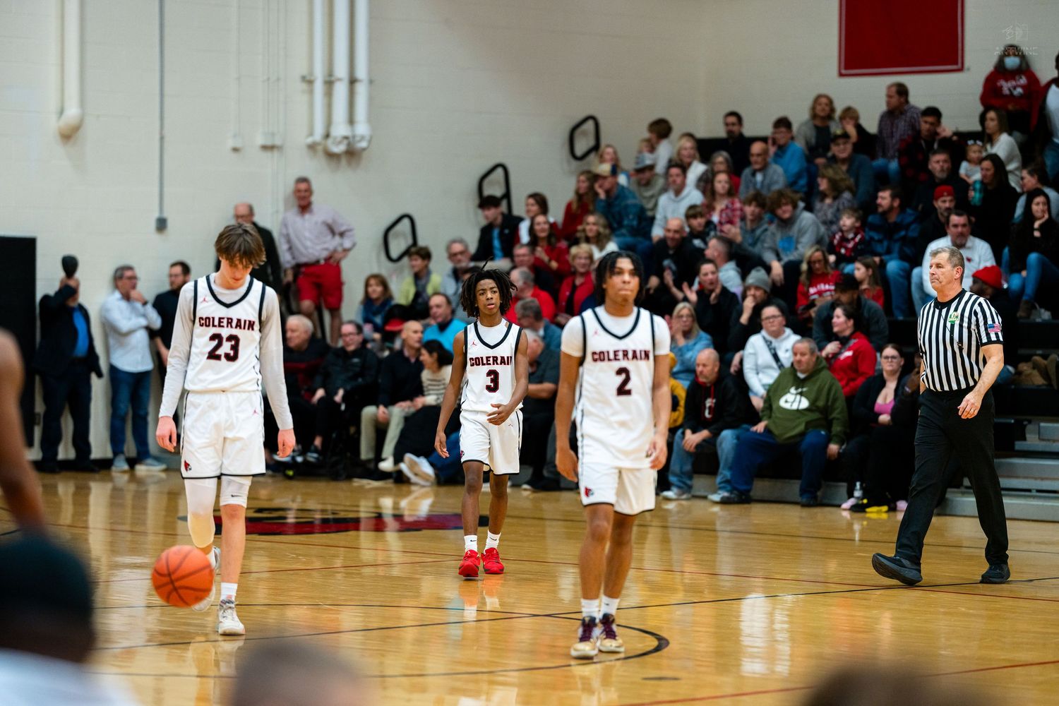 Three Cardinals basketball players walk on court during high school game with packed stands.