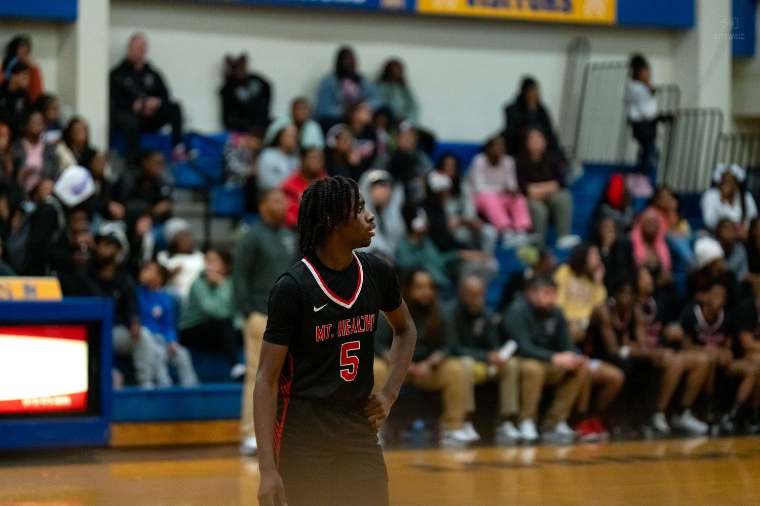 Basketball player #5 in black Mt. Healthy uniform stands on the court with fans in the background.