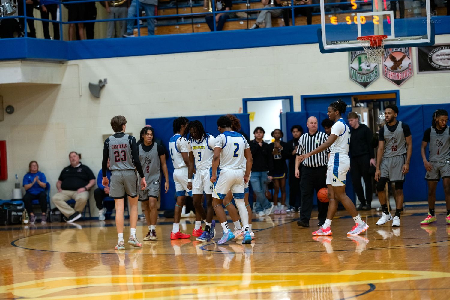 High school basketball players and referee gather on the court during a timeout in a blue and white gymnasium.