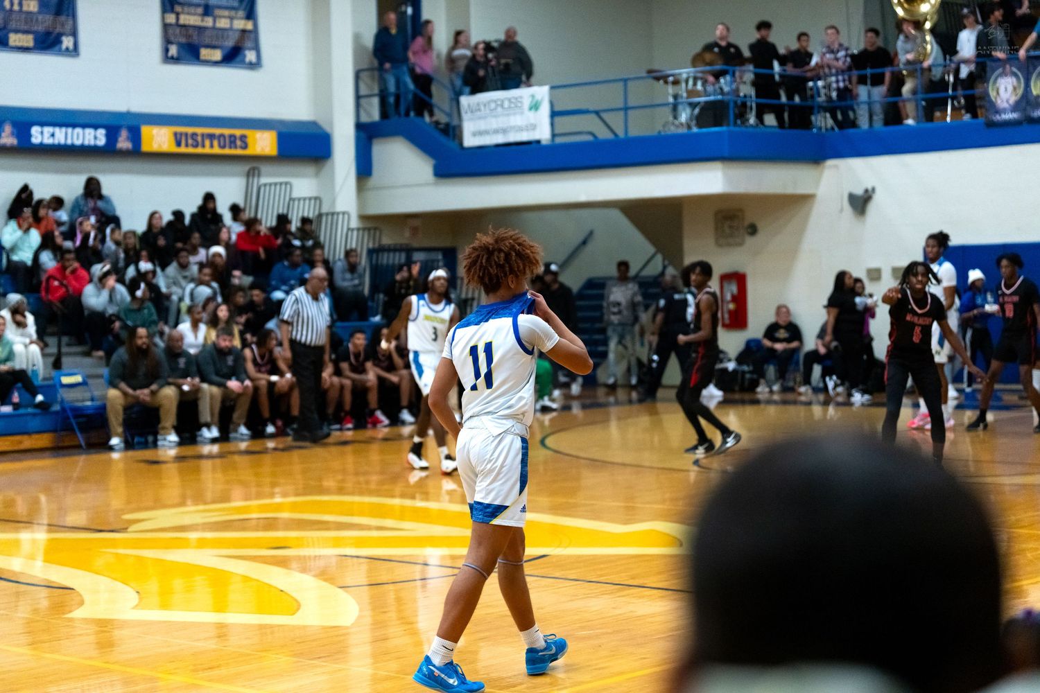 Basketball player wearing number 11 white jersey walks on court during packed high school game.
