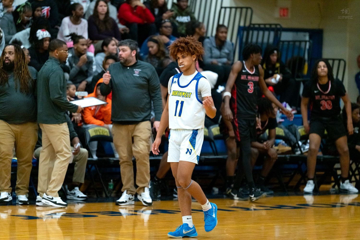 Player #11 in blue uniform walks on basketball court during competitive high school game with crowded bleachers.