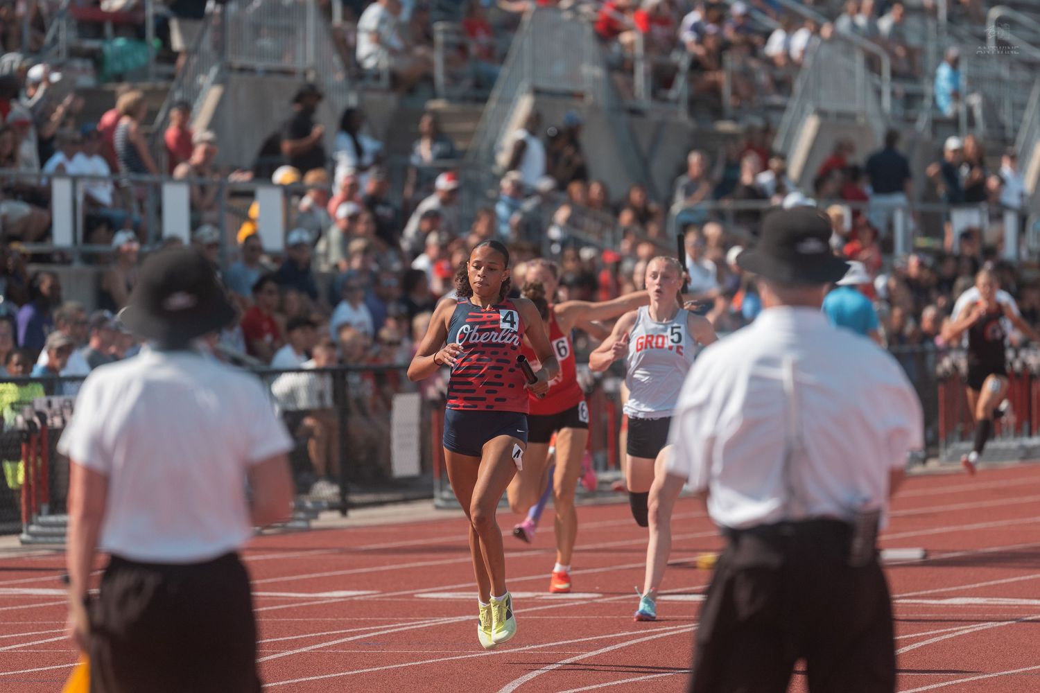Female track athletes sprint past officials on a red track at a competitive outdoor meet.