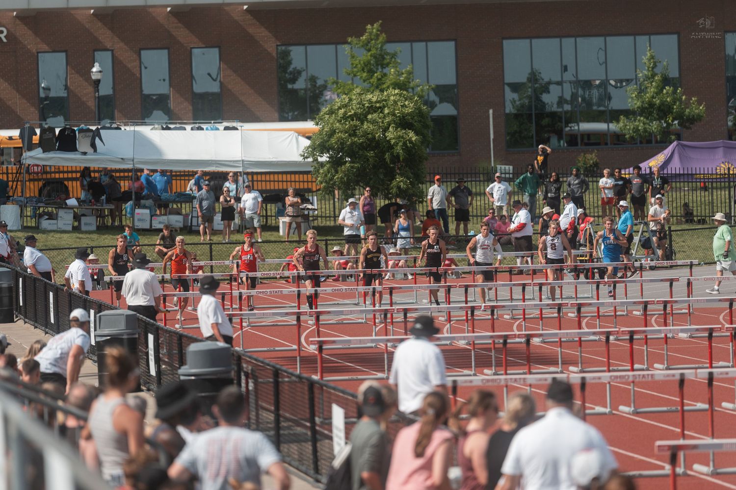 Athletes competing in hurdles event on red track with school bus and building visible in background.