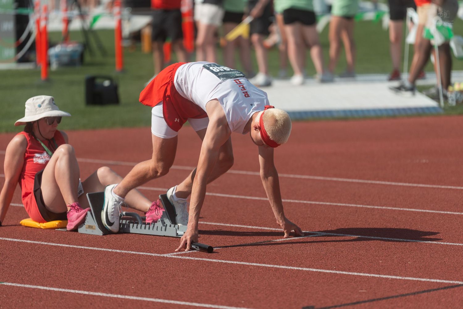 Senior athlete in white uniform prepares in starting blocks on red track at outdoor competition.