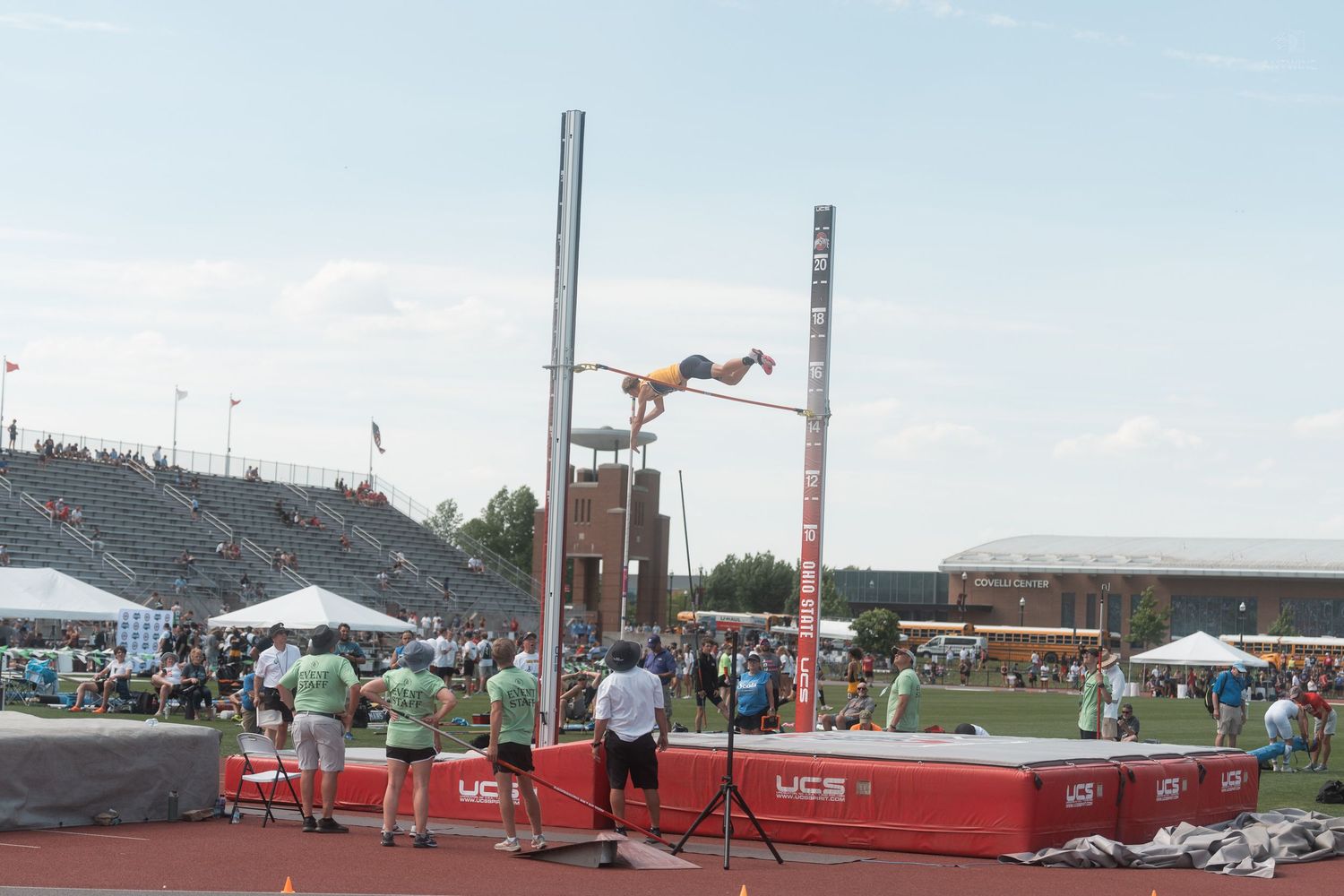 Pole vaulter clears the bar in orange uniform at a competitive outdoor track and field championship event.