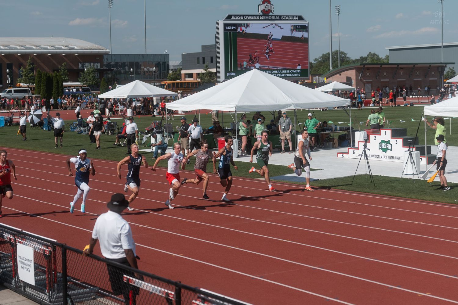 Athletes race on a red outdoor track during a competitive track and field meet with spectators lining the course.