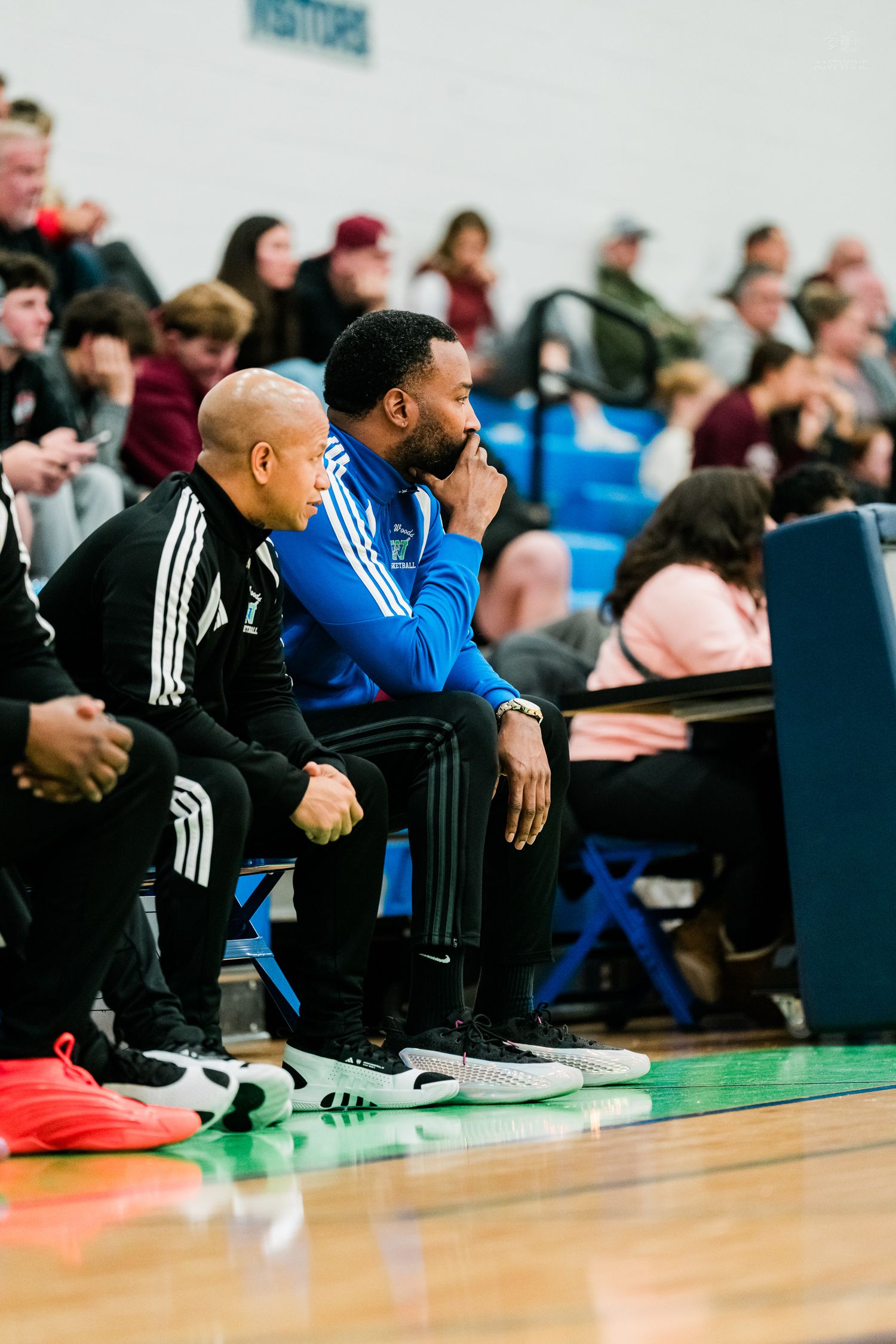 Two coaches in black and blue athletic wear sit on the bench observing a high school basketball game.