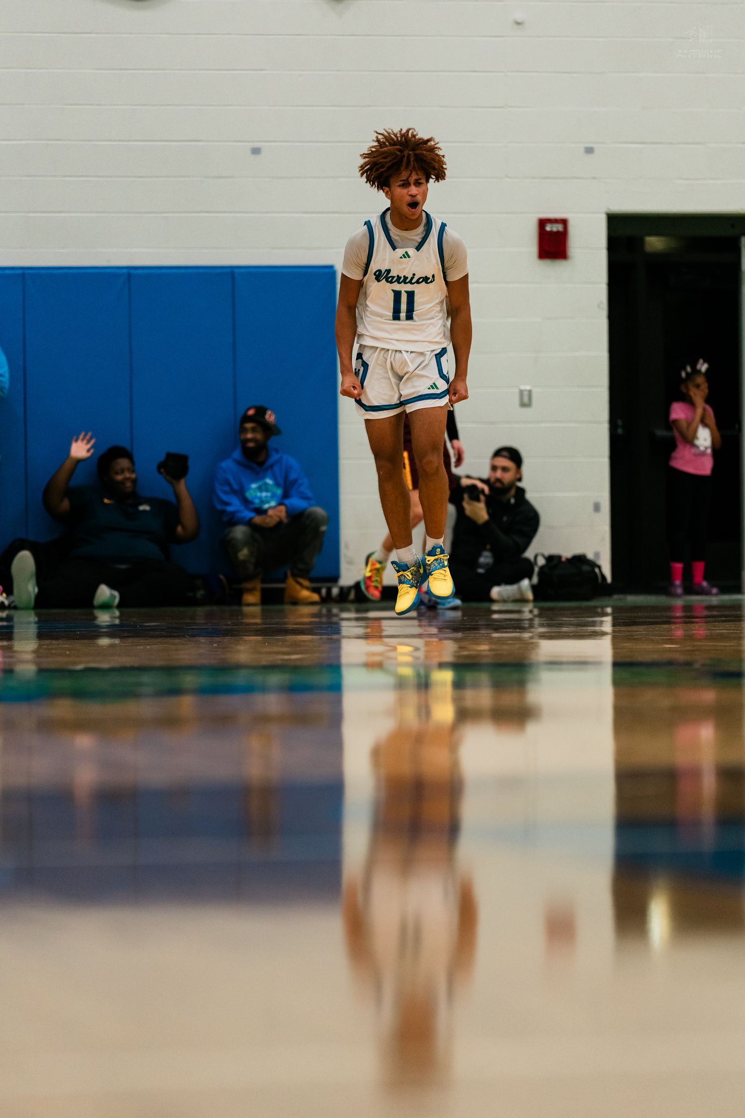 High school basketball player wearing number 11 white Warrior uniform jumps on reflective court with fans watching.