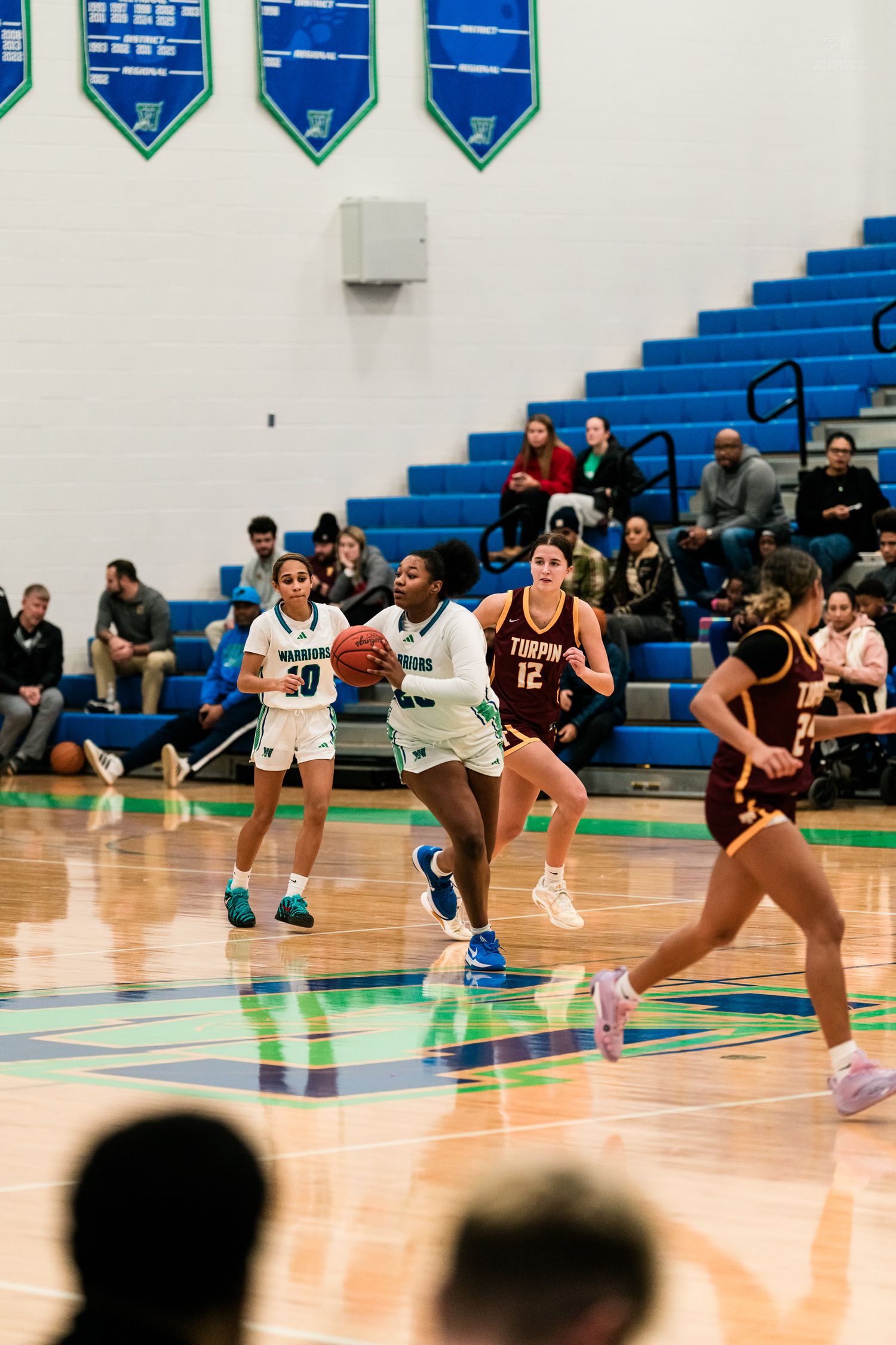 High school girls basketball player in teal uniform drives toward the basket against maroon-uniformed defenders in a blue gym.