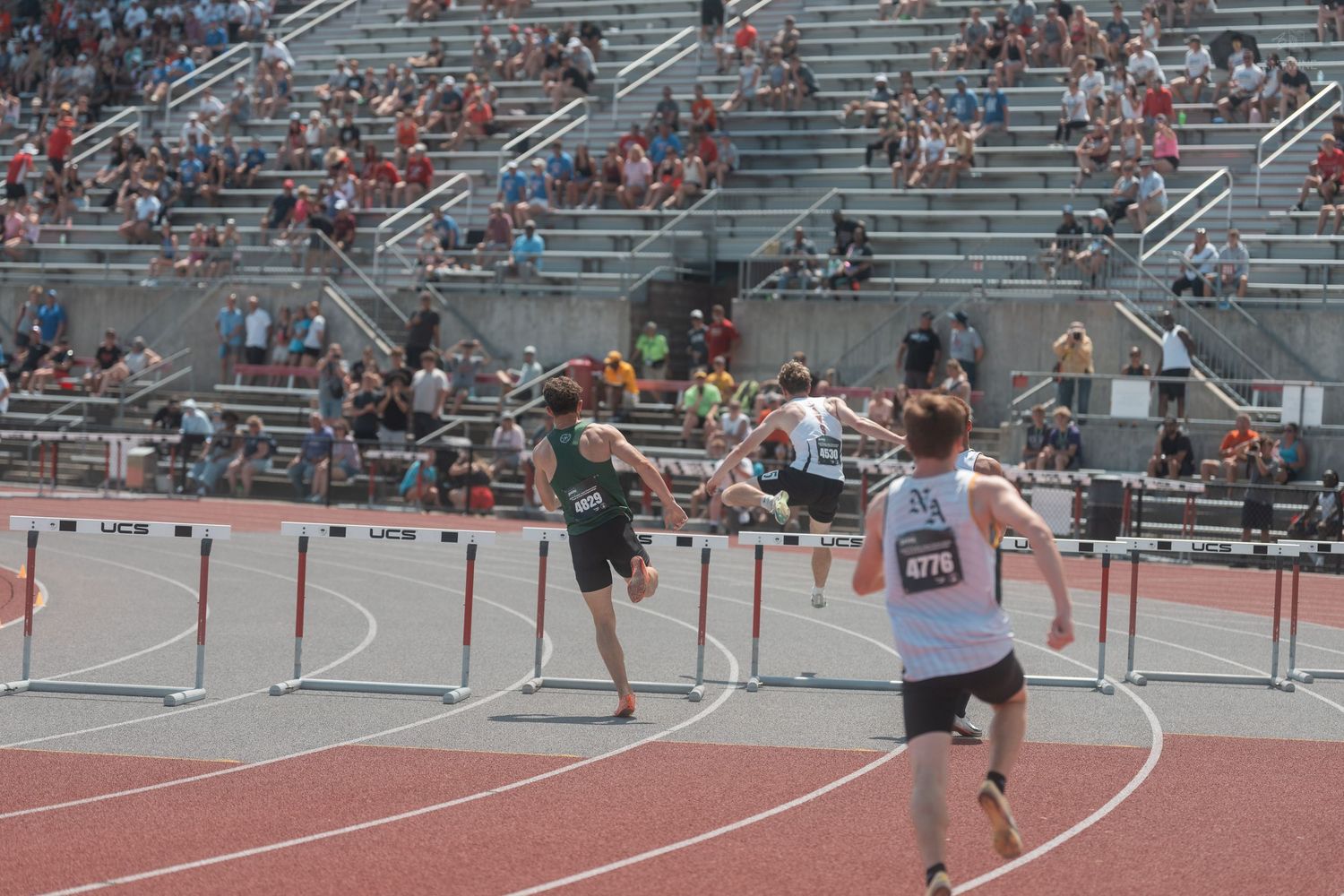 Runners clear hurdles during a track meet with packed bleachers of spectators in the background.