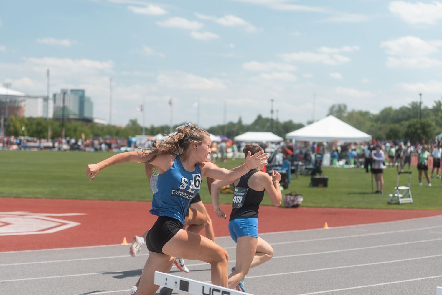 Two female athletes clearing a hurdle simultaneously during an outdoor track and field competition on a sunny day.