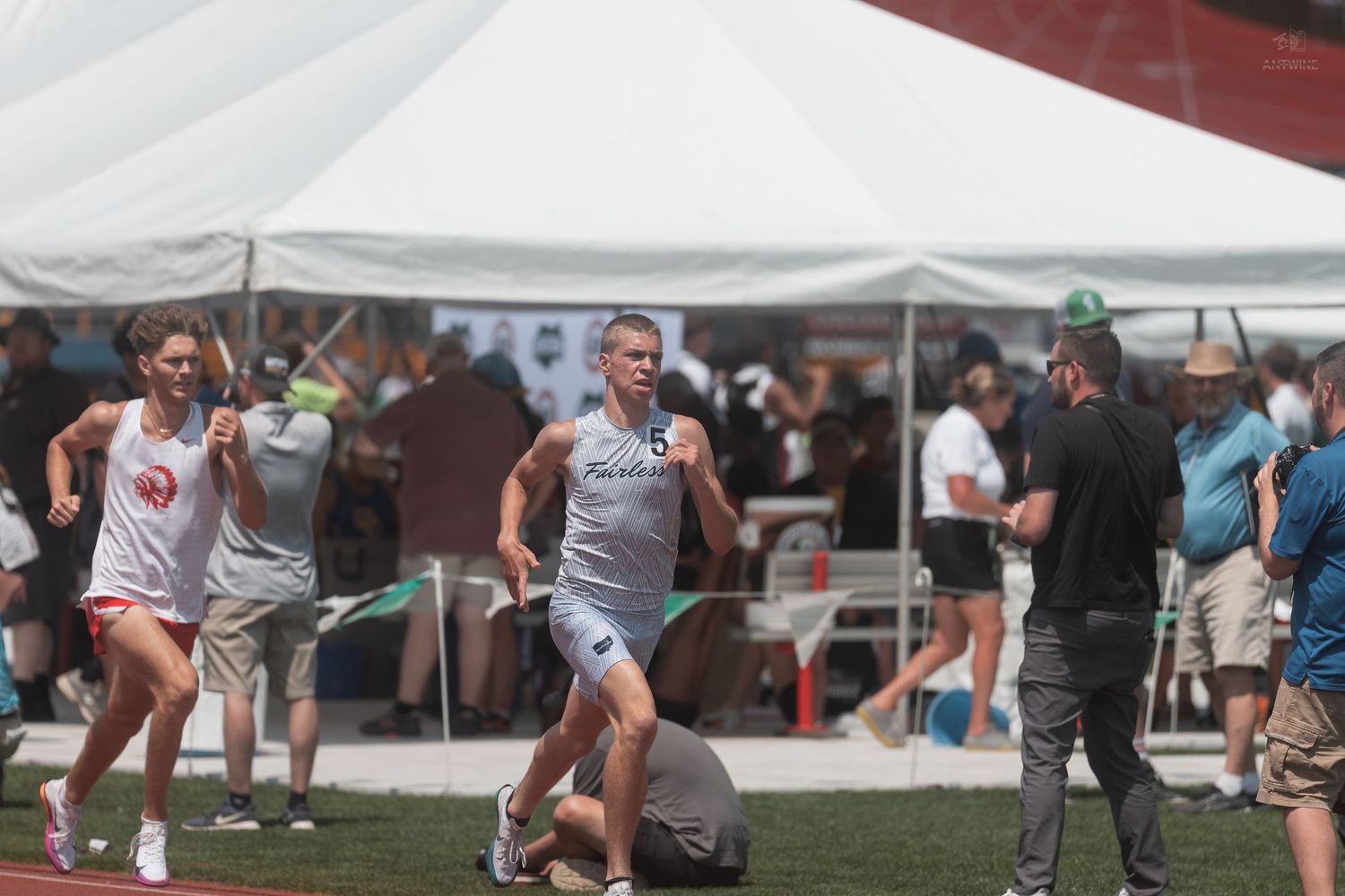 Two runners compete in a track race near a white tent at an outdoor athletic competition.