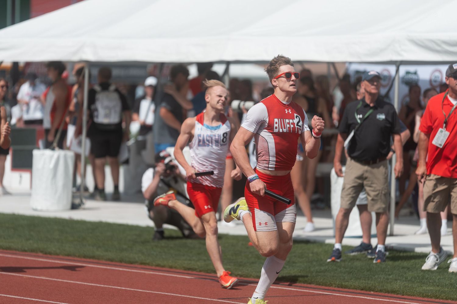 A Clippers athlete leads competitors during a relay race at a sunny outdoor track and field championship.