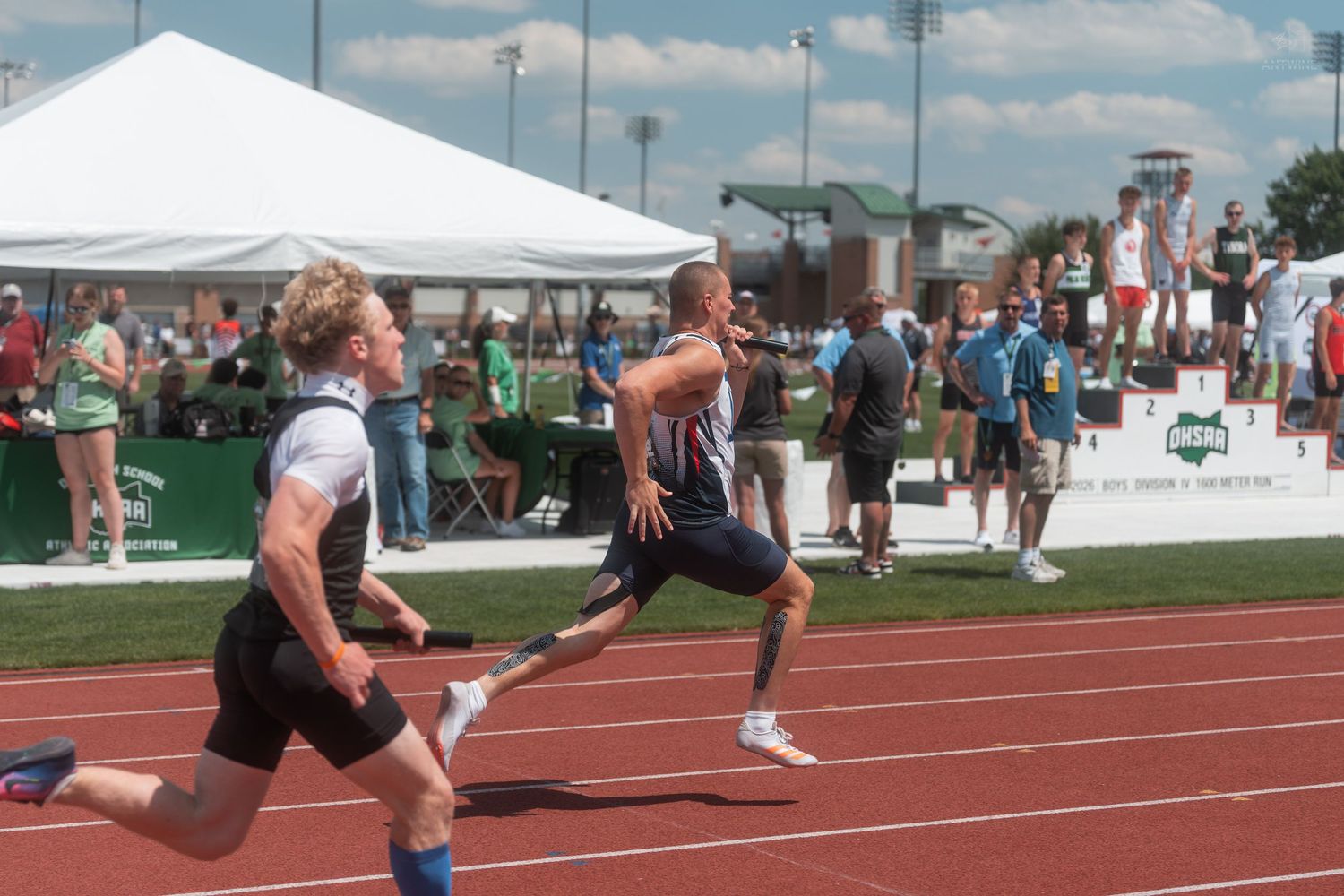 Two male sprinters race on a red track at an outdoor athletic competition with spectators watching from the sidelines.
