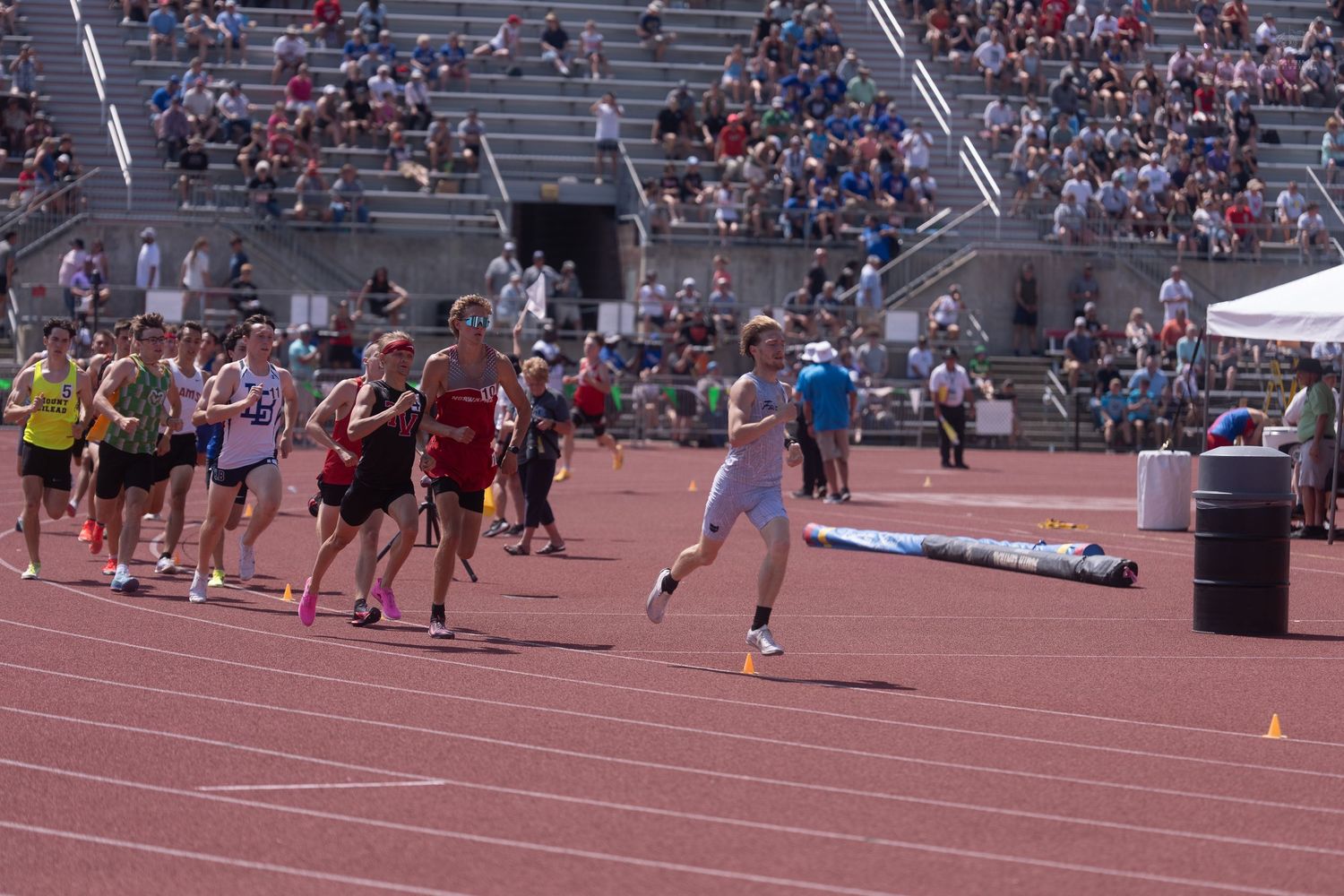 Runners compete in a track relay race passing the baton in front of packed stadium bleachers.