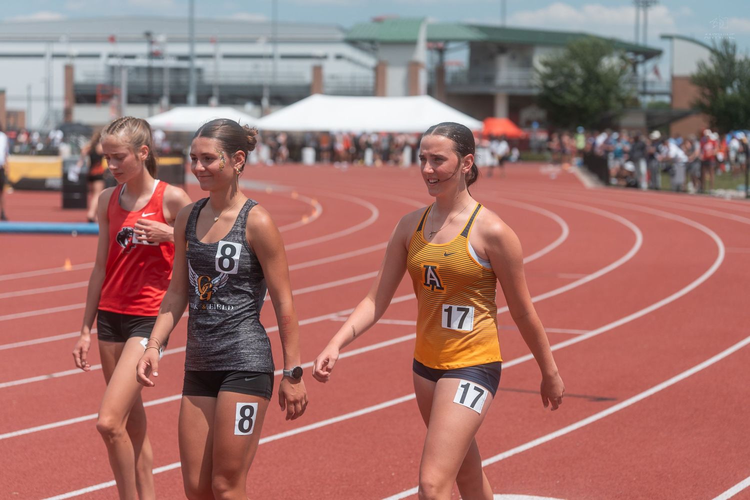 Three female track athletes in numbered bibs walking on red track after completing outdoor race event.
