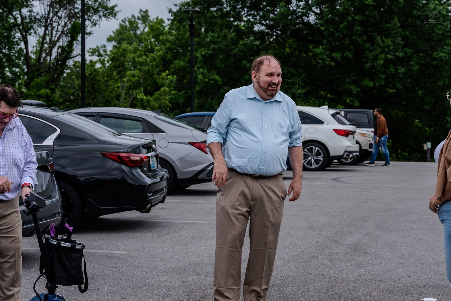 Heavyset man in light blue shirt standing in outdoor parking lot with cars and green trees.