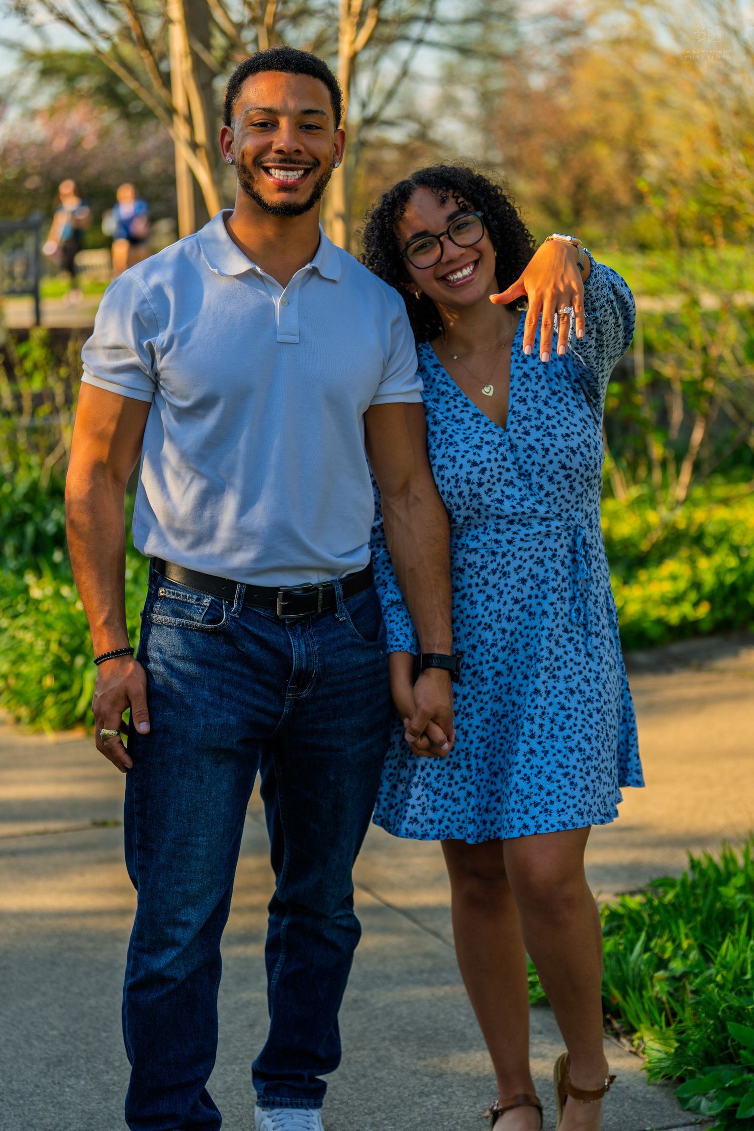 A joyful couple smiles and embraces on a sunny park path, the woman showing off her engagement ring.