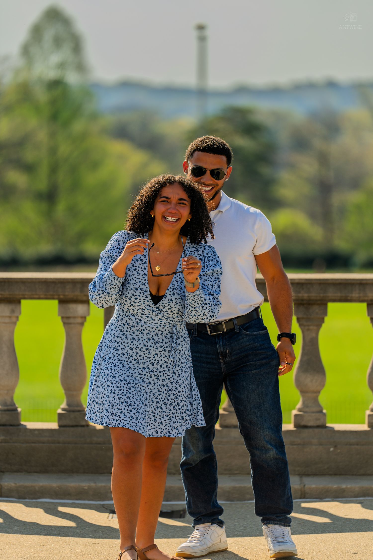 A happy couple poses together on a stone balcony with a scenic green park landscape behind them.