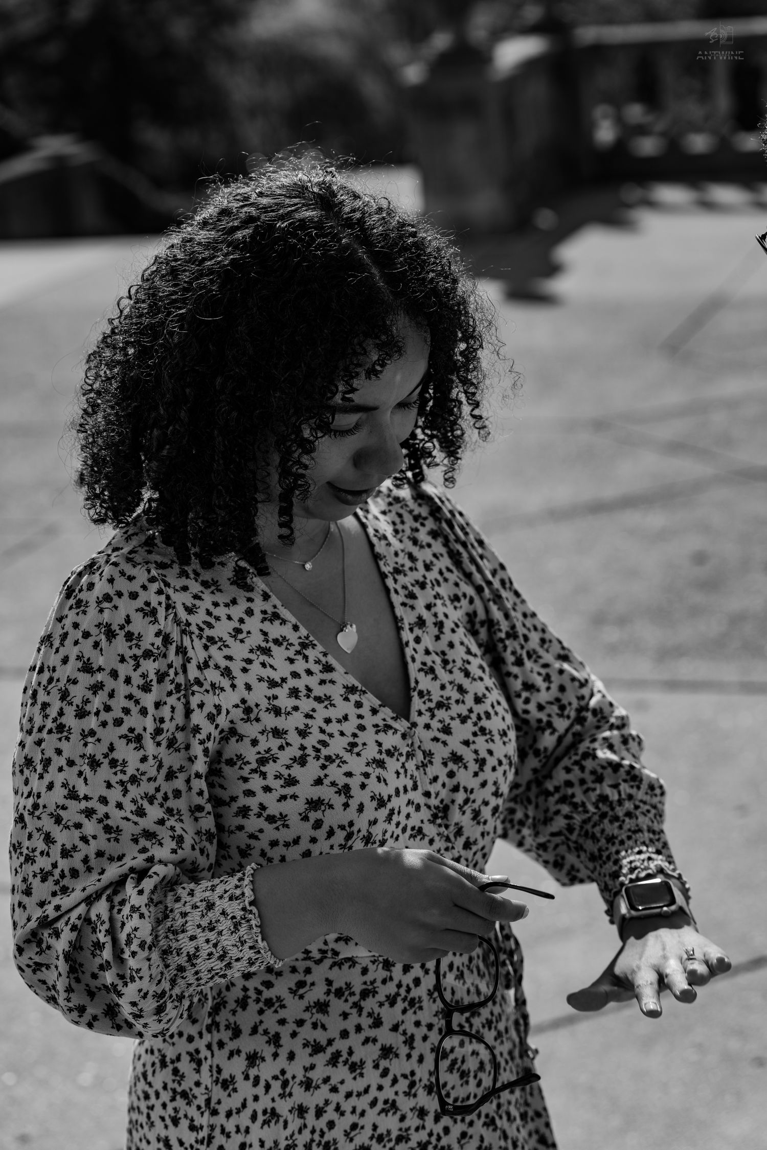 Black and white portrait of a curly-haired woman in a floral dress holding a phone and sunglasses outdoors.