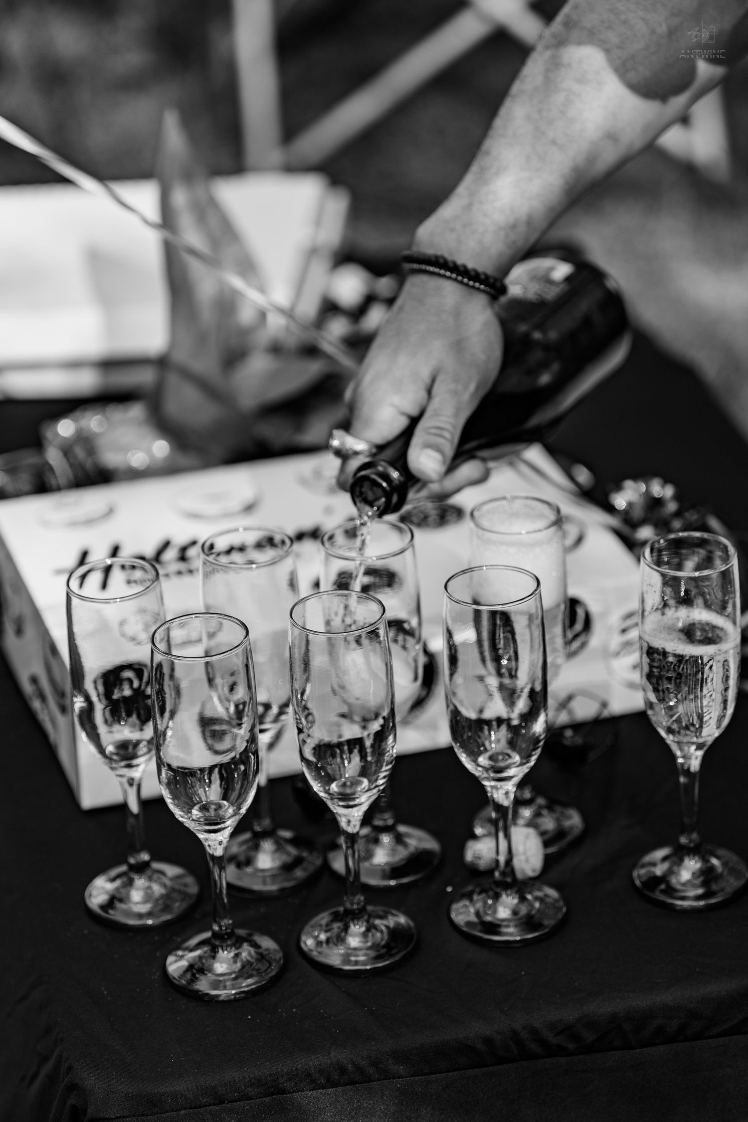 Black and white photo of hand pouring champagne into elegant crystal flutes at outdoor event.