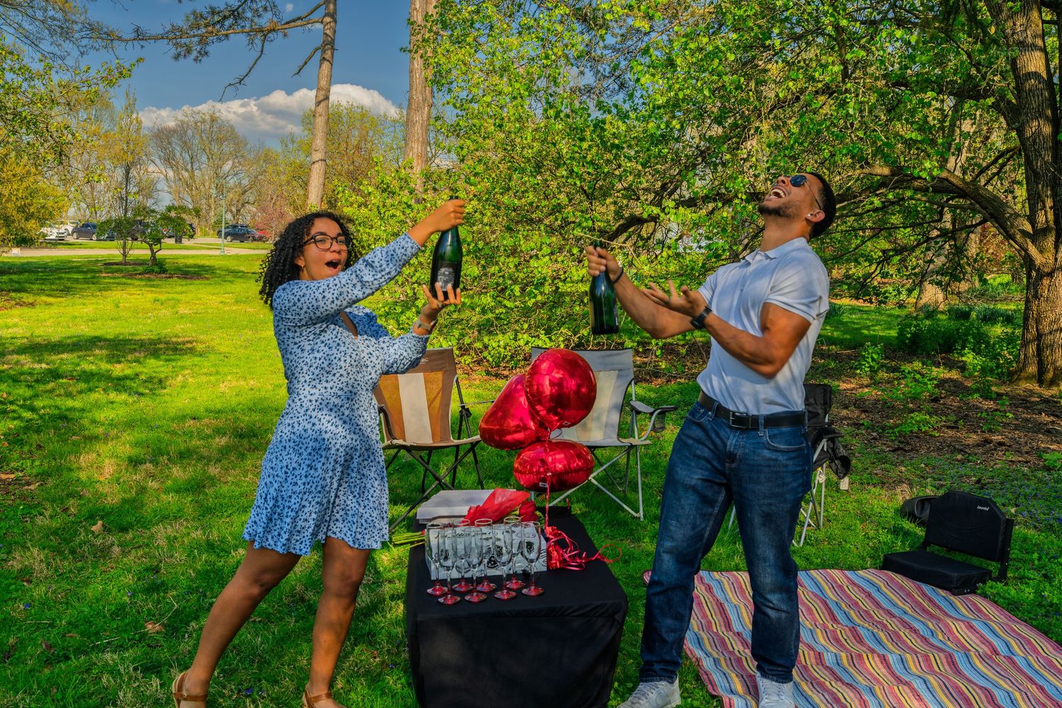 Joyful couple raising champagne bottles outdoors surrounded by red heart balloons during a sunny park picnic.