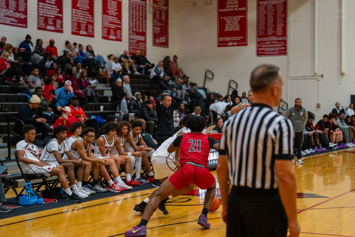 A basketball player in red dribbles near the sideline as a referee watches during a high school game.