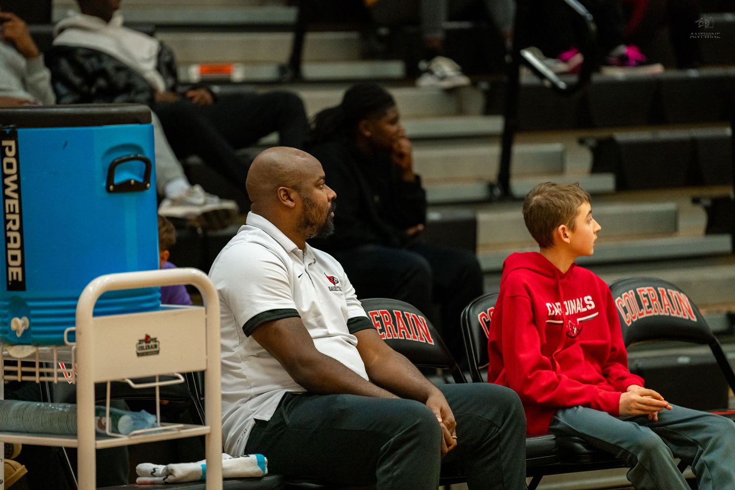 A coach and young fan sit courtside watching a high school basketball game near a Powerade cooler.