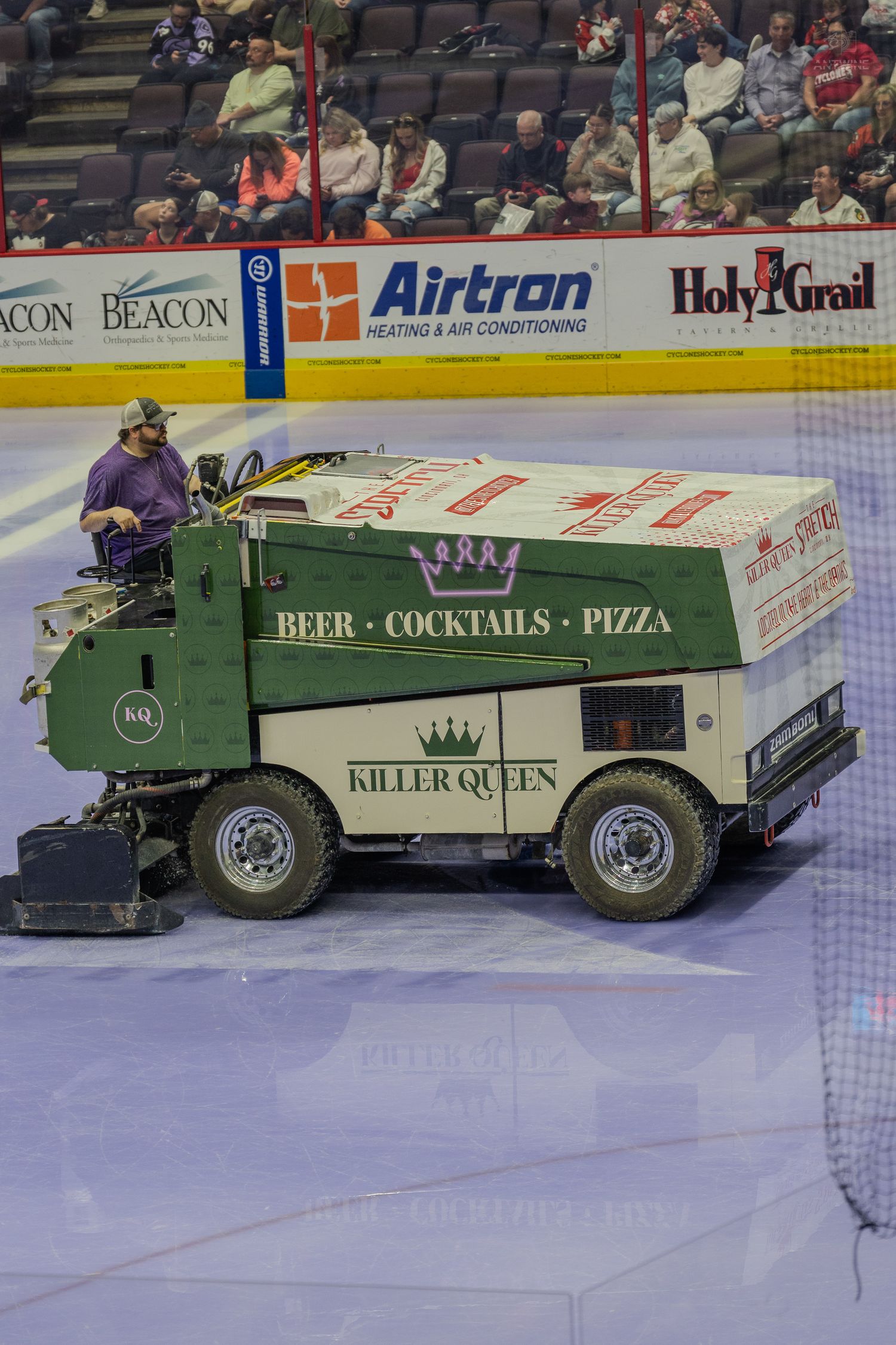 A green Killer Queen branded Zamboni advertising beer, cocktails, and pizza resurfacing the ice at a hockey arena.