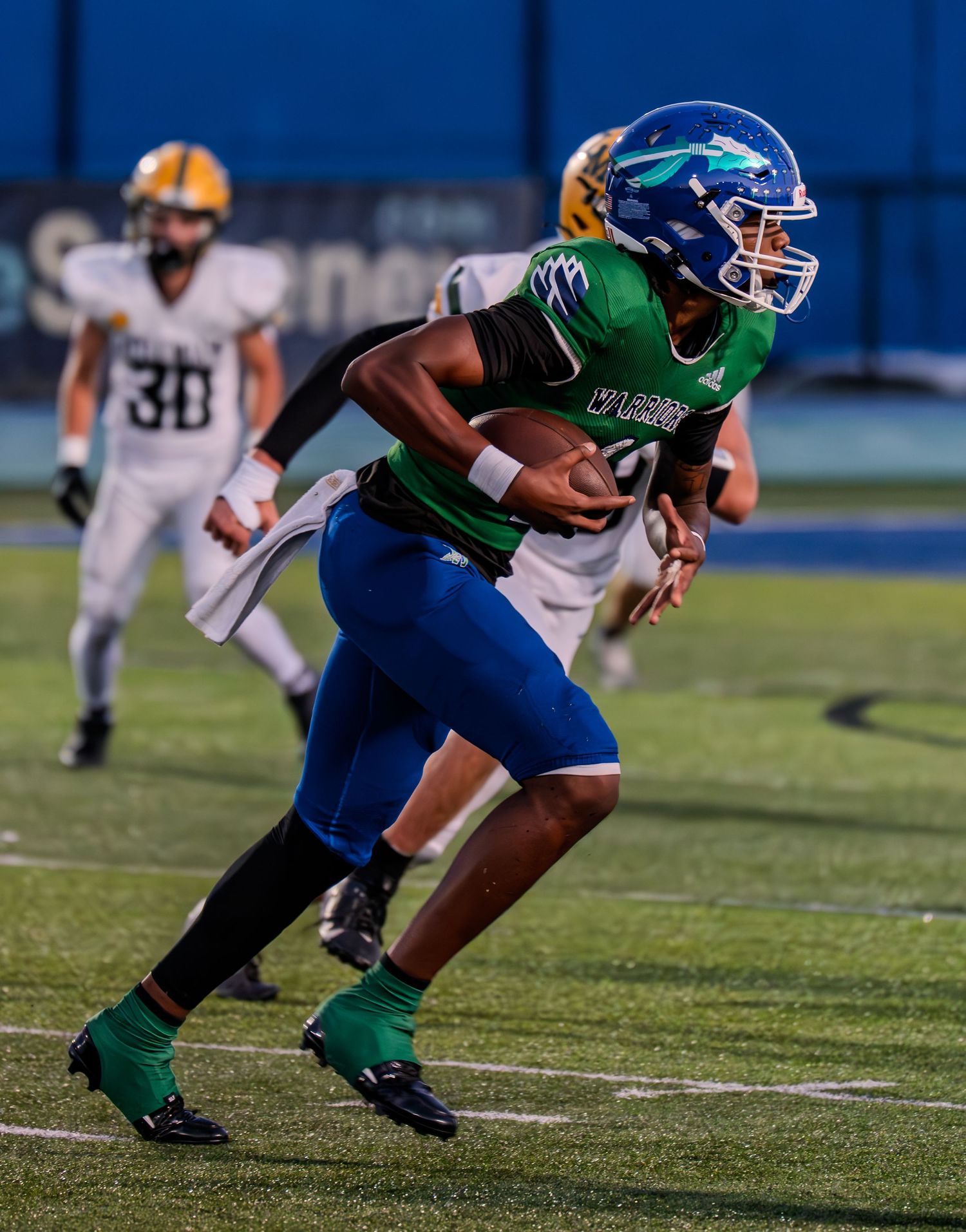 A Warriors ball carrier in green jersey runs with the football during a night game under stadium lights.