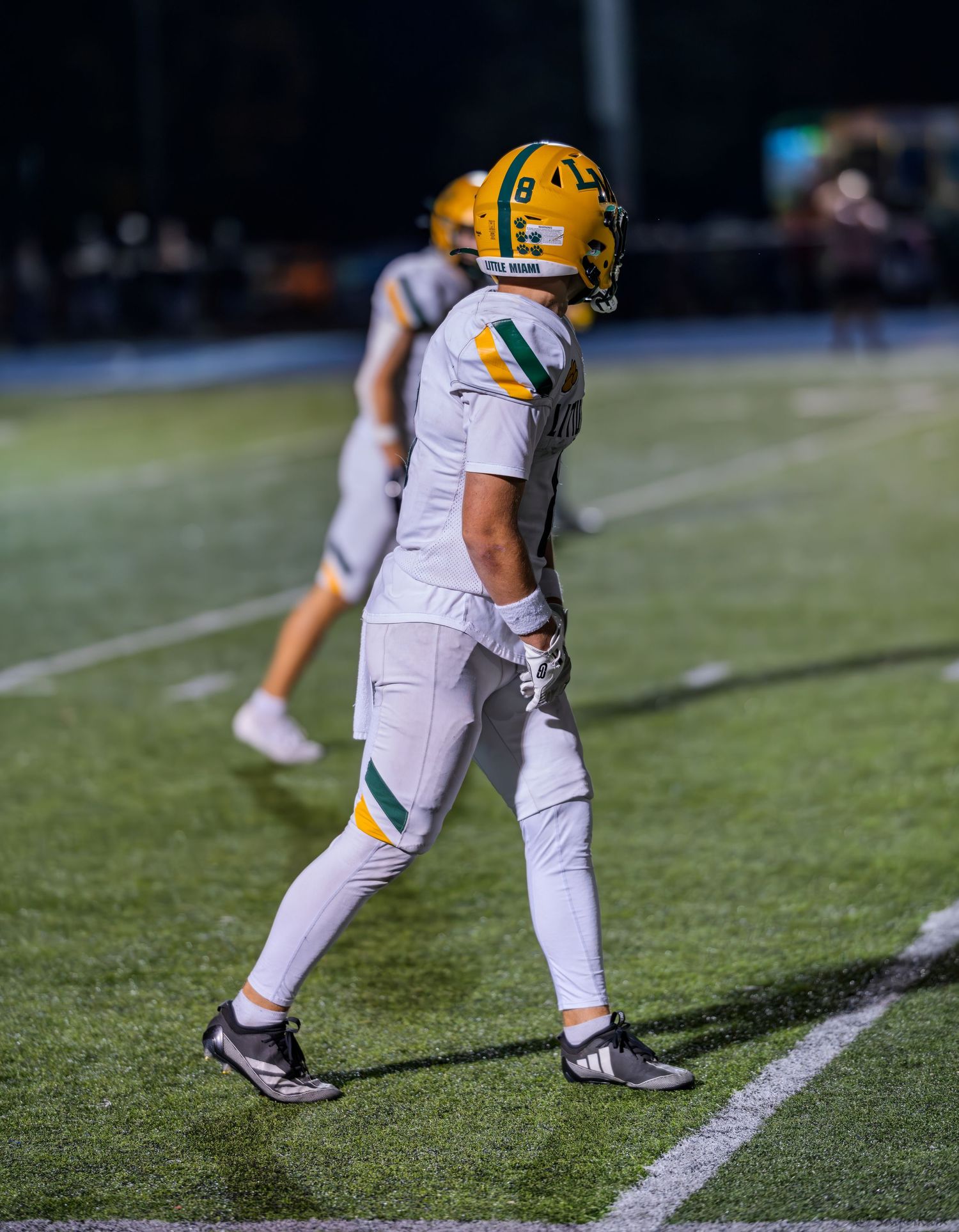 A football player in white and gold uniform walks along the sideline during a night game.