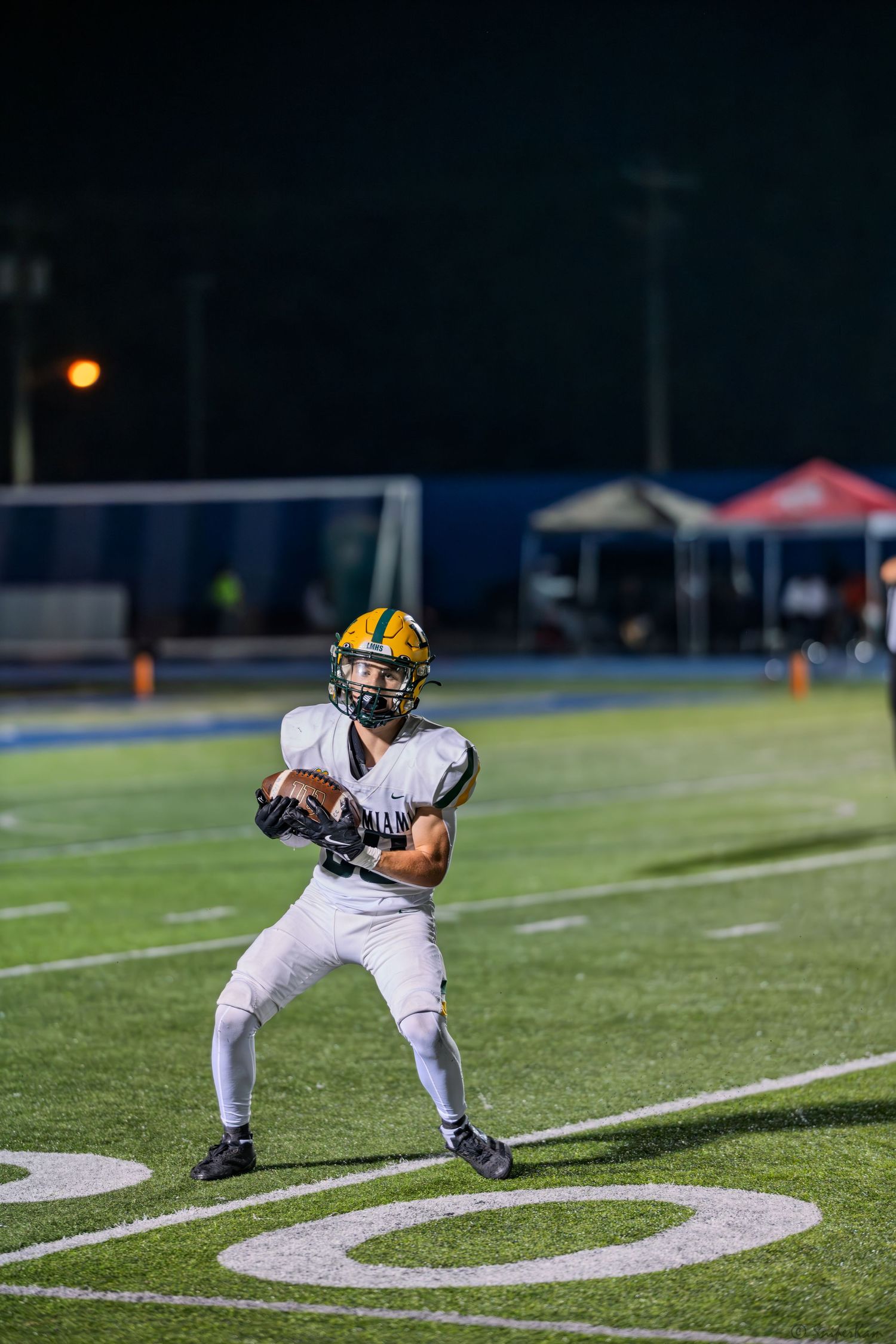 High school football player in white and gold uniform catches a pass near the sideline at night.