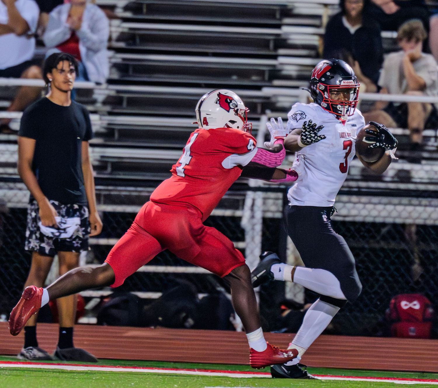 High school football receiver in white uniform catches a pass while being defended during a night game.