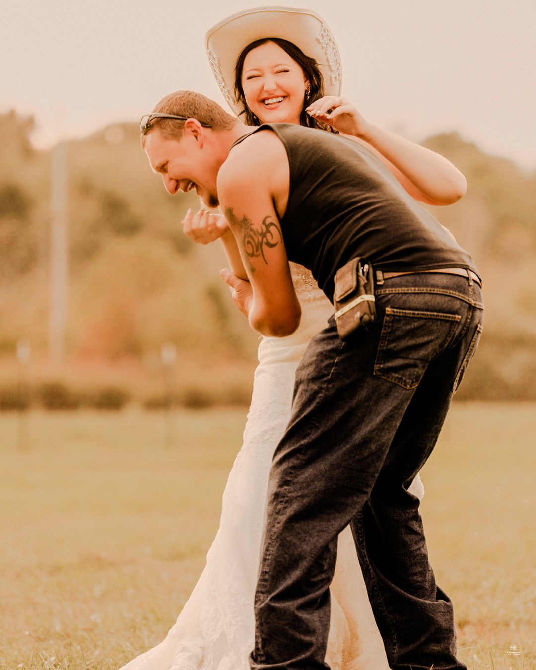 Laughing bride in white dress and cowboy hat playfully jumping on grooms back at outdoor wedding.