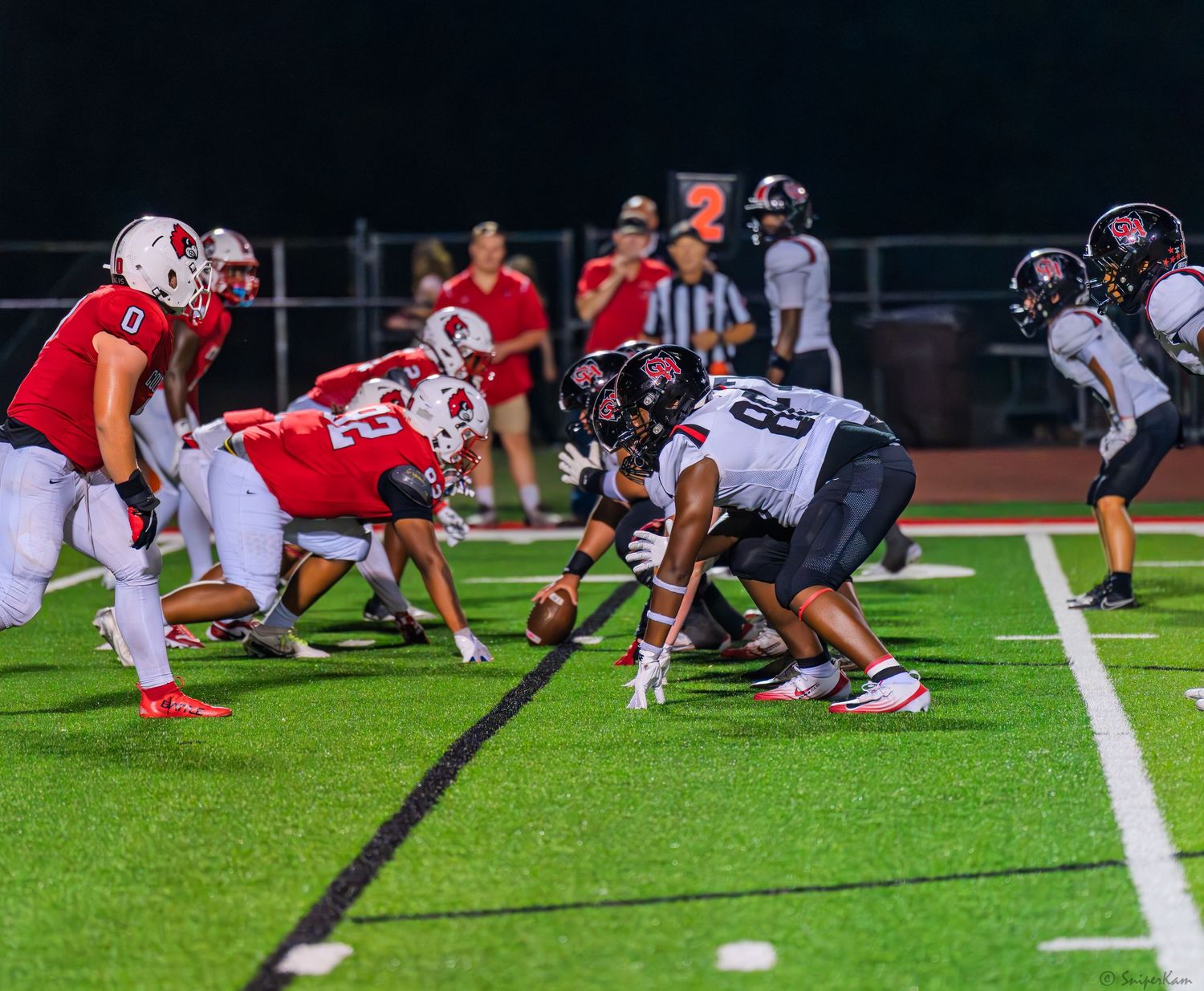High school football teams line up at the line of scrimmage during a night game on green turf.