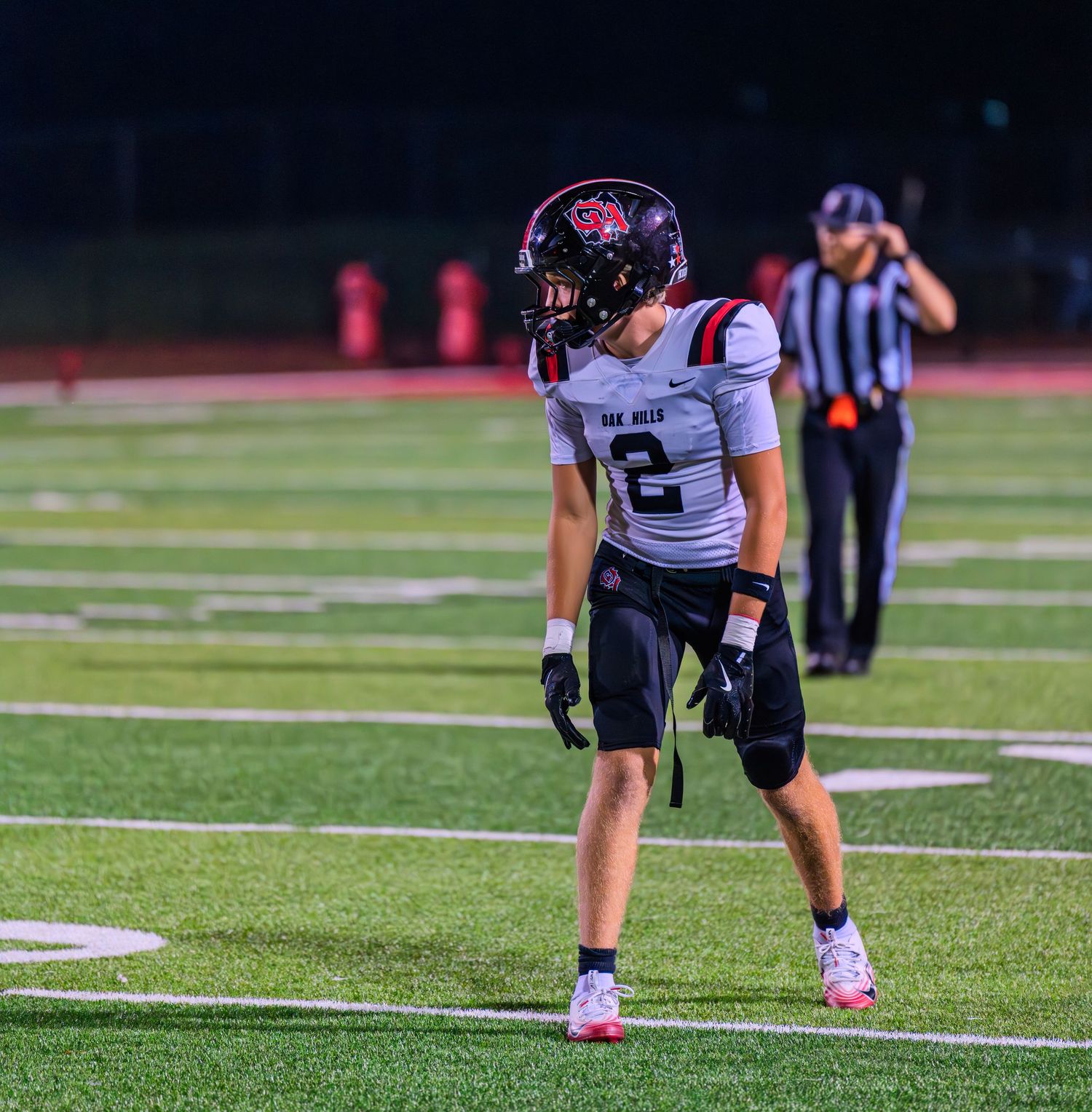 High school football player #2 in white Oak Hills jersey stands in ready position on the field during a night game.