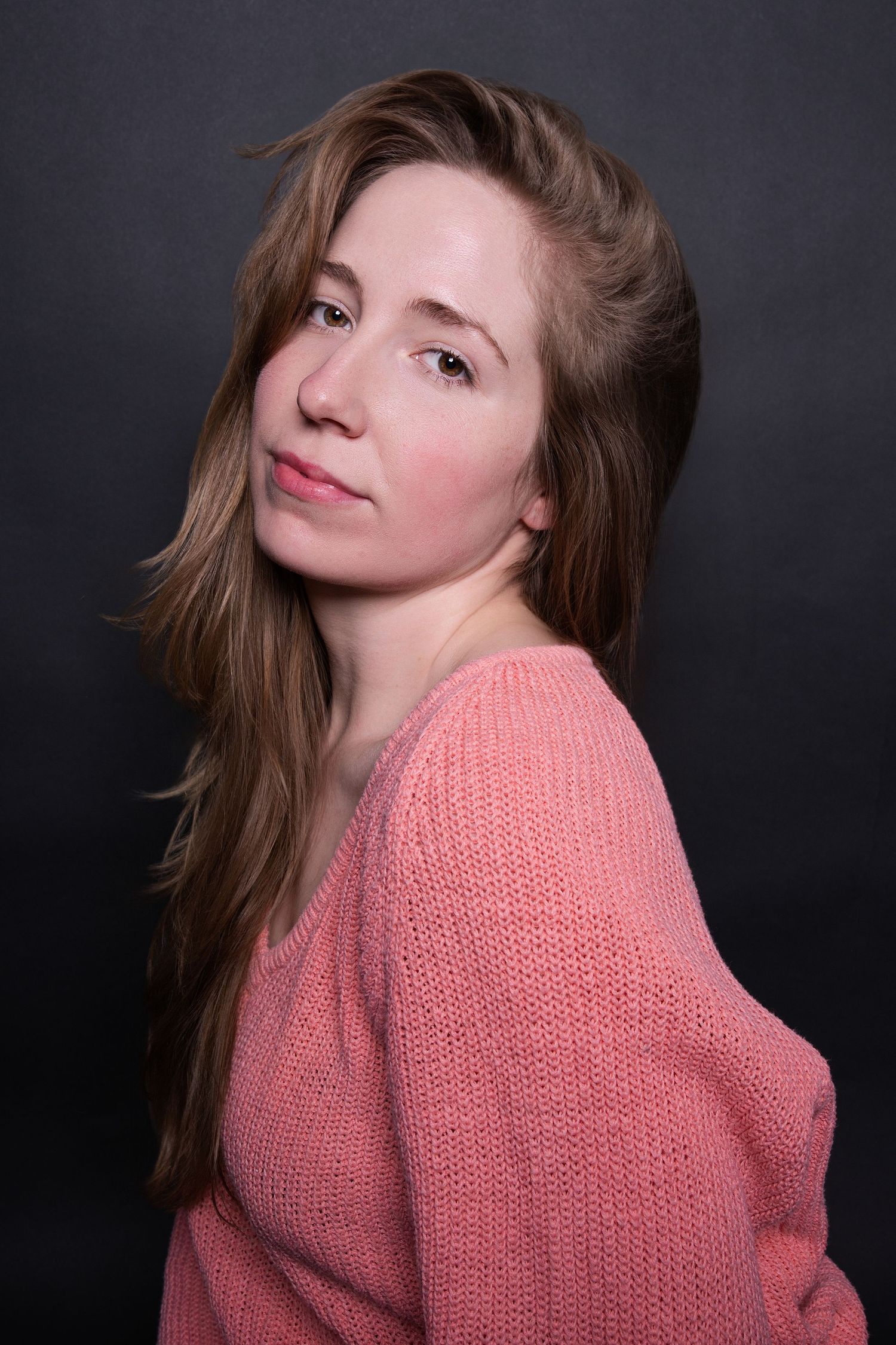Woman with long brown hair in a pink knit sweater glancing back over her shoulder in a studio.